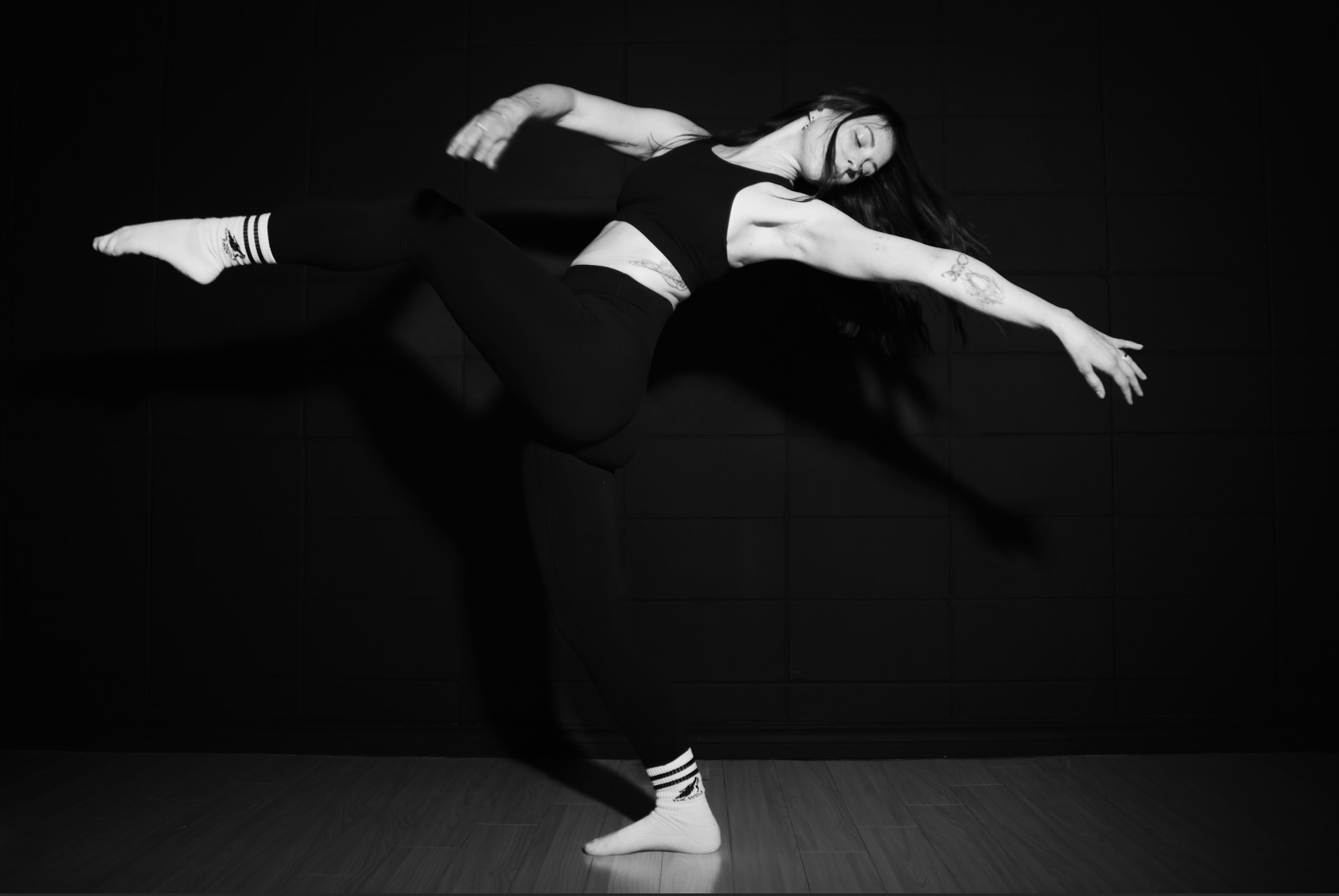 A woman in black athletic wear performing a dance or fitness move against a dark background.