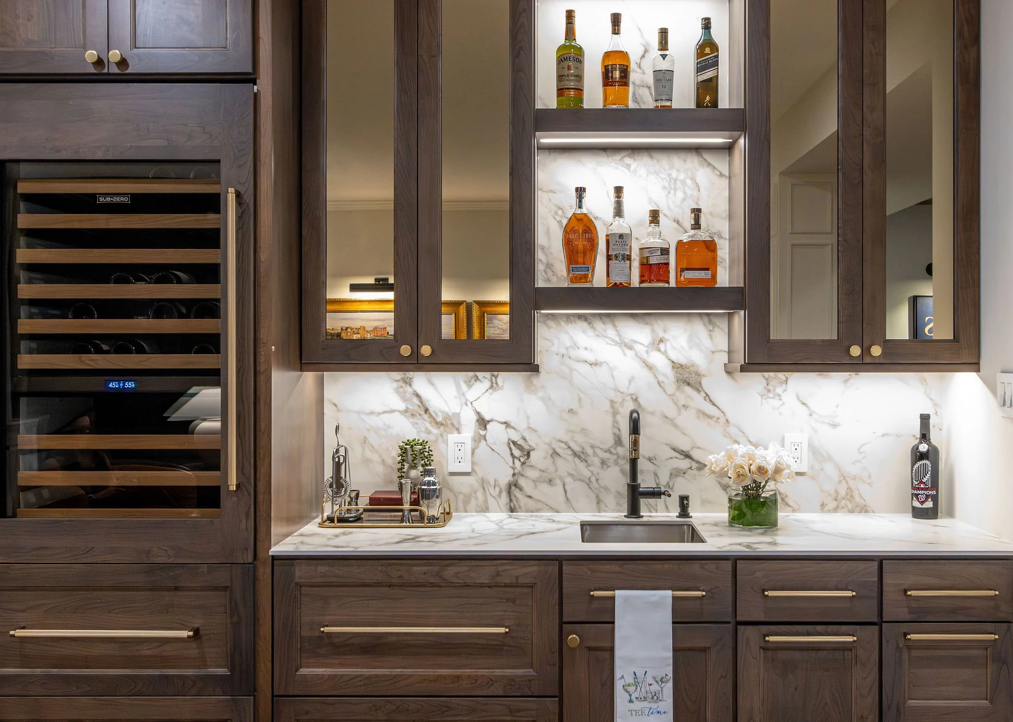 bar with cherry cabinets, marble backsplash, wine fridge on the left, open shelves with liquor bottles, sink with black faucet, flowers in a vase, and soap dispenser.