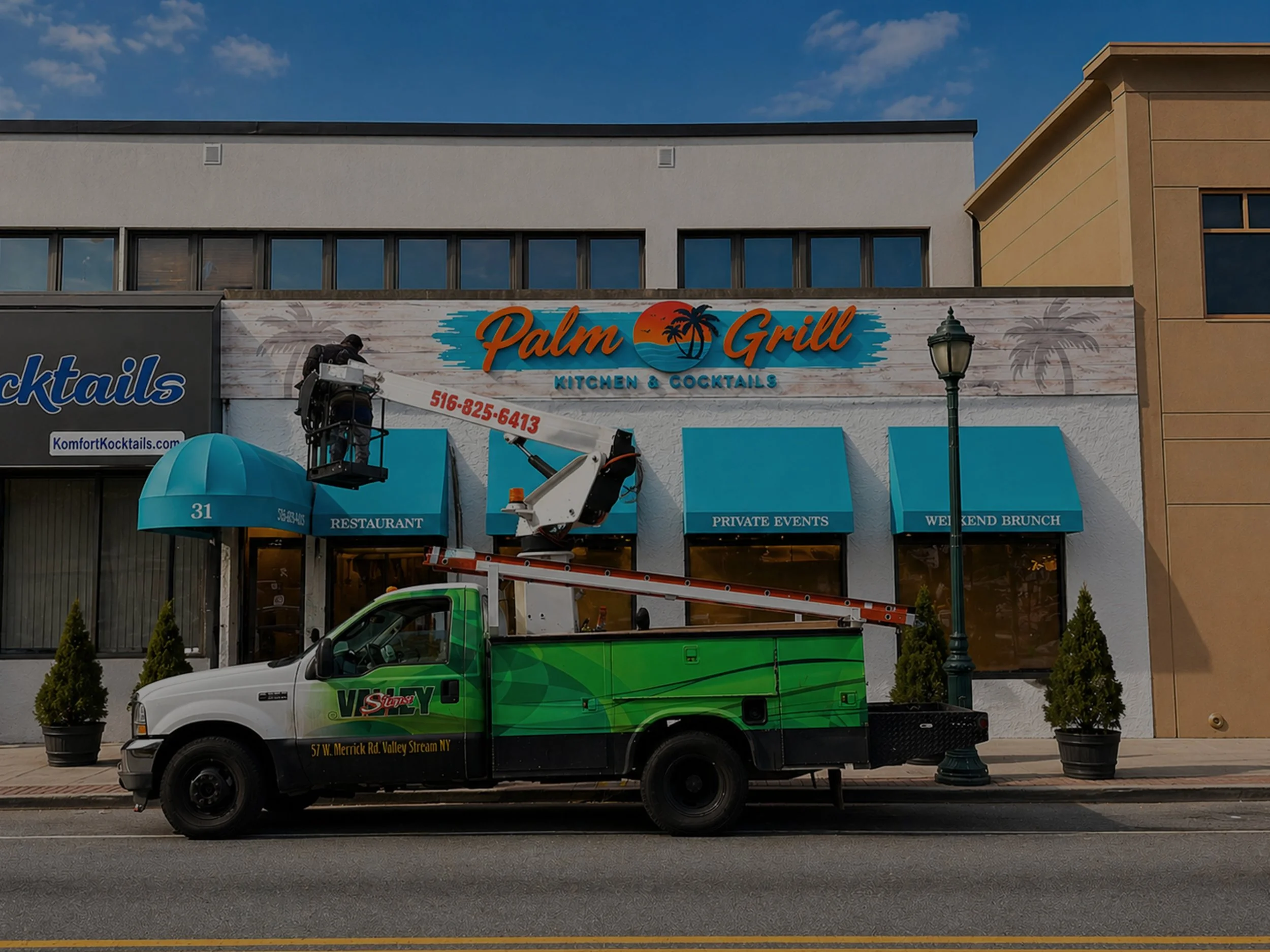 People installing a scoreboard on a brick building using a cherry picker with a green and white utility truck parked nearby.