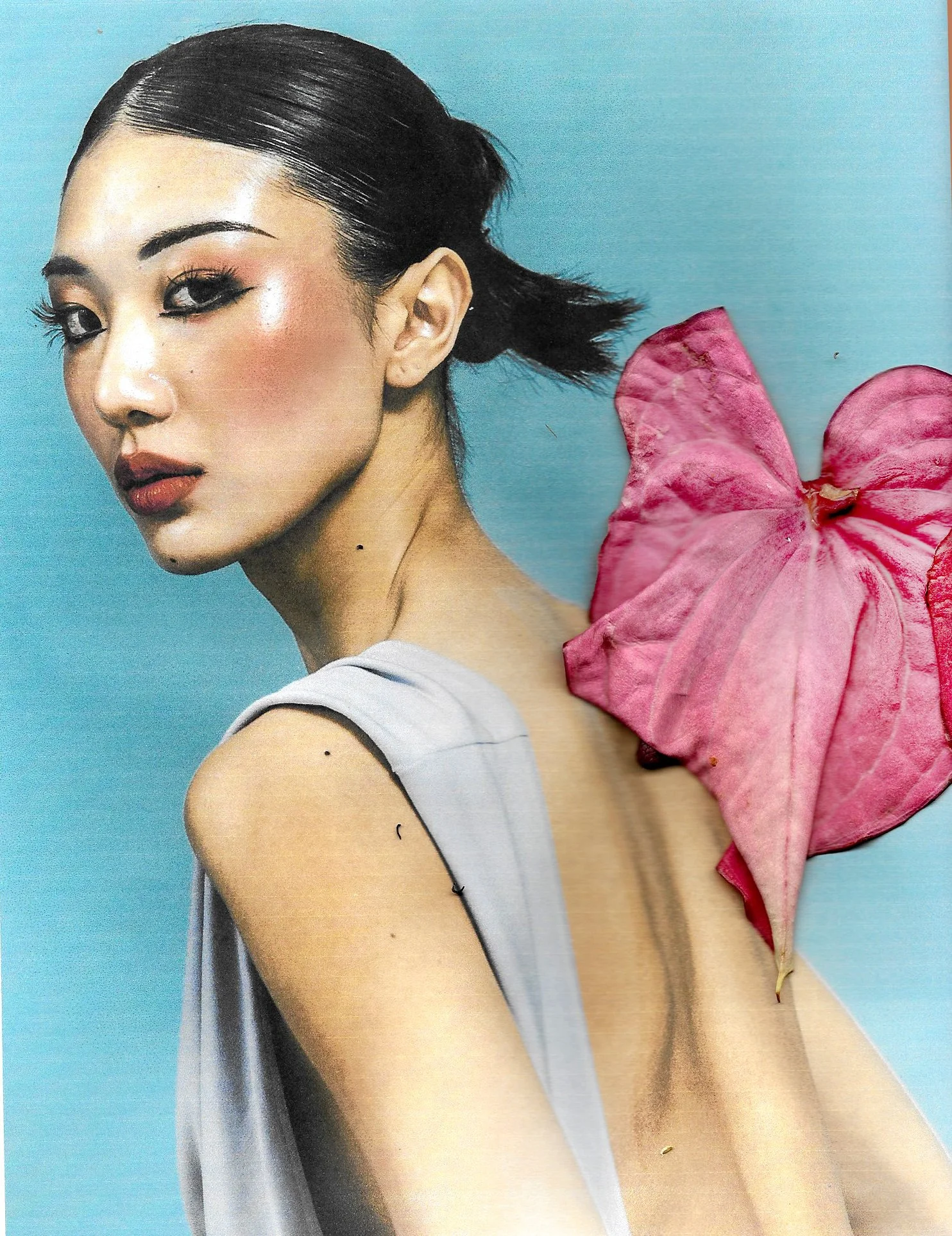 A woman with dark hair pulled back, wearing makeup, with a pink flower on her shoulder, against a light blue background.