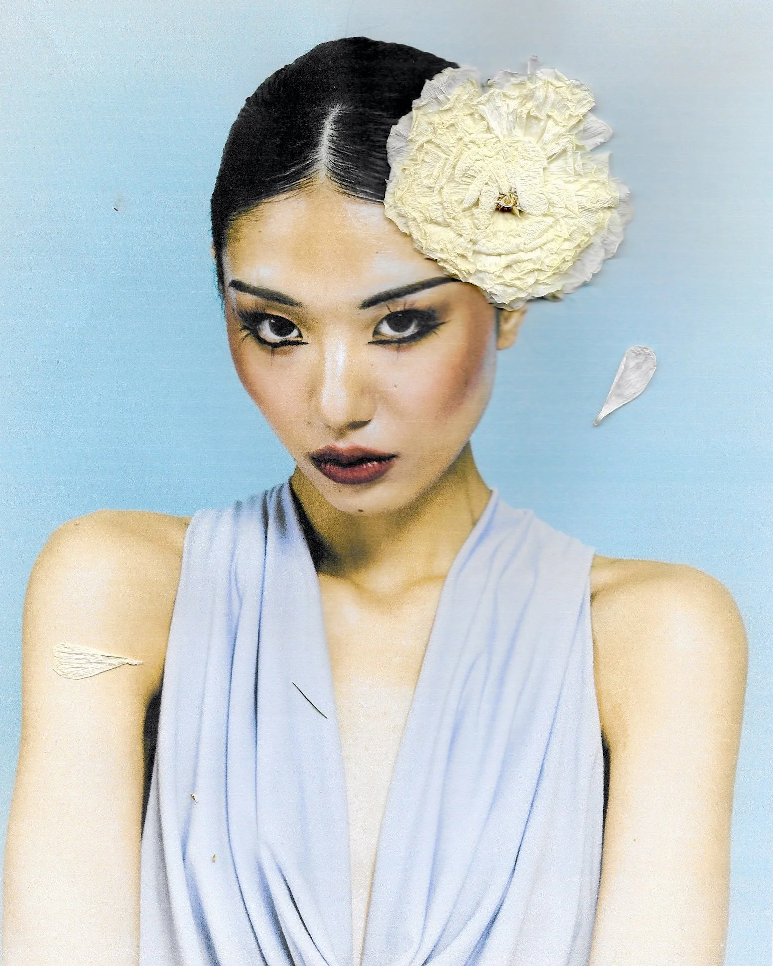 A woman with dark hair styled back, wearing a light blue dress, with makeup, and a large cream-colored flower in her hair, against a light blue background.