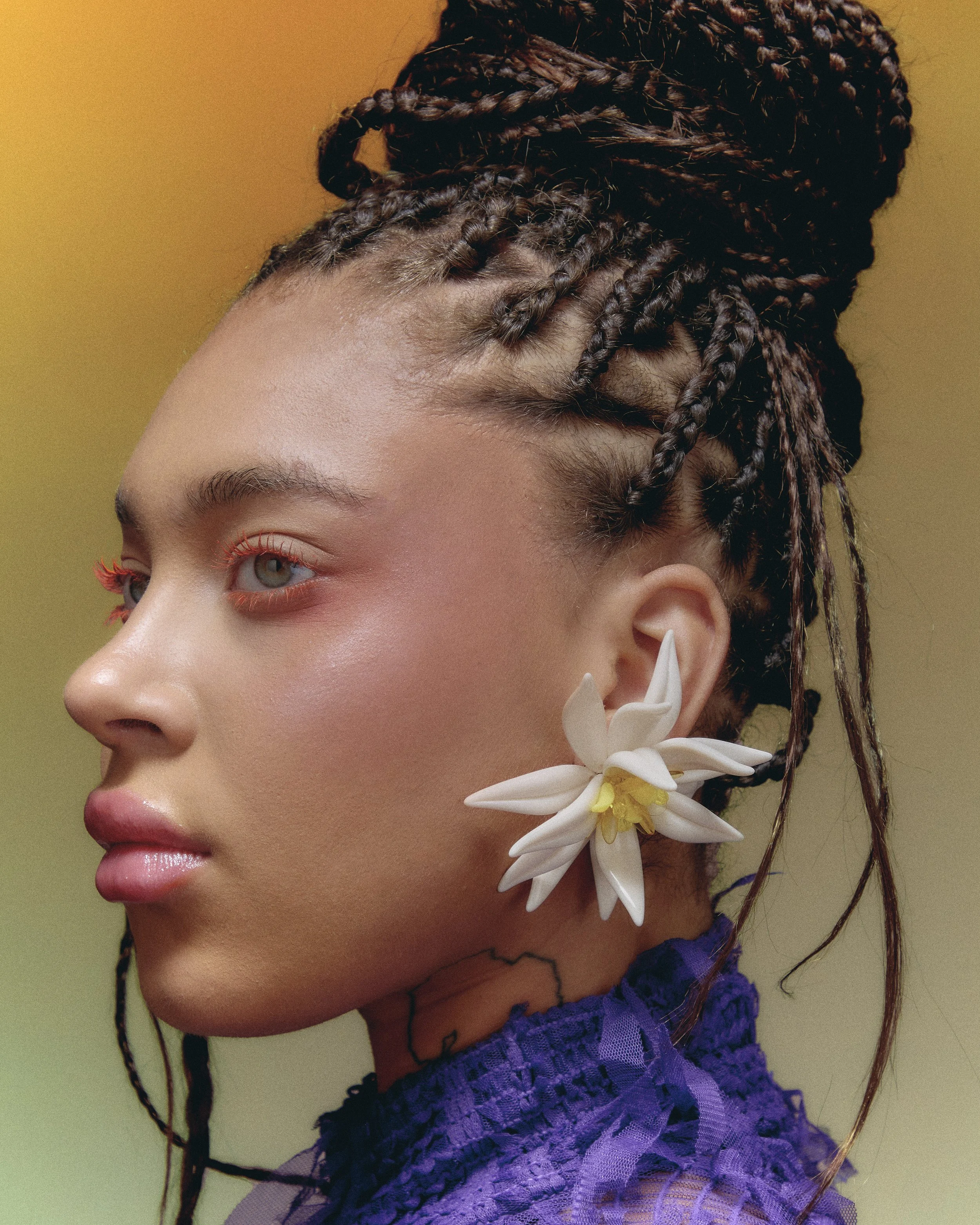 A woman with braided hair and a large white flower earring, wearing a purple lace top, showcasing a side profile against a yellow background.