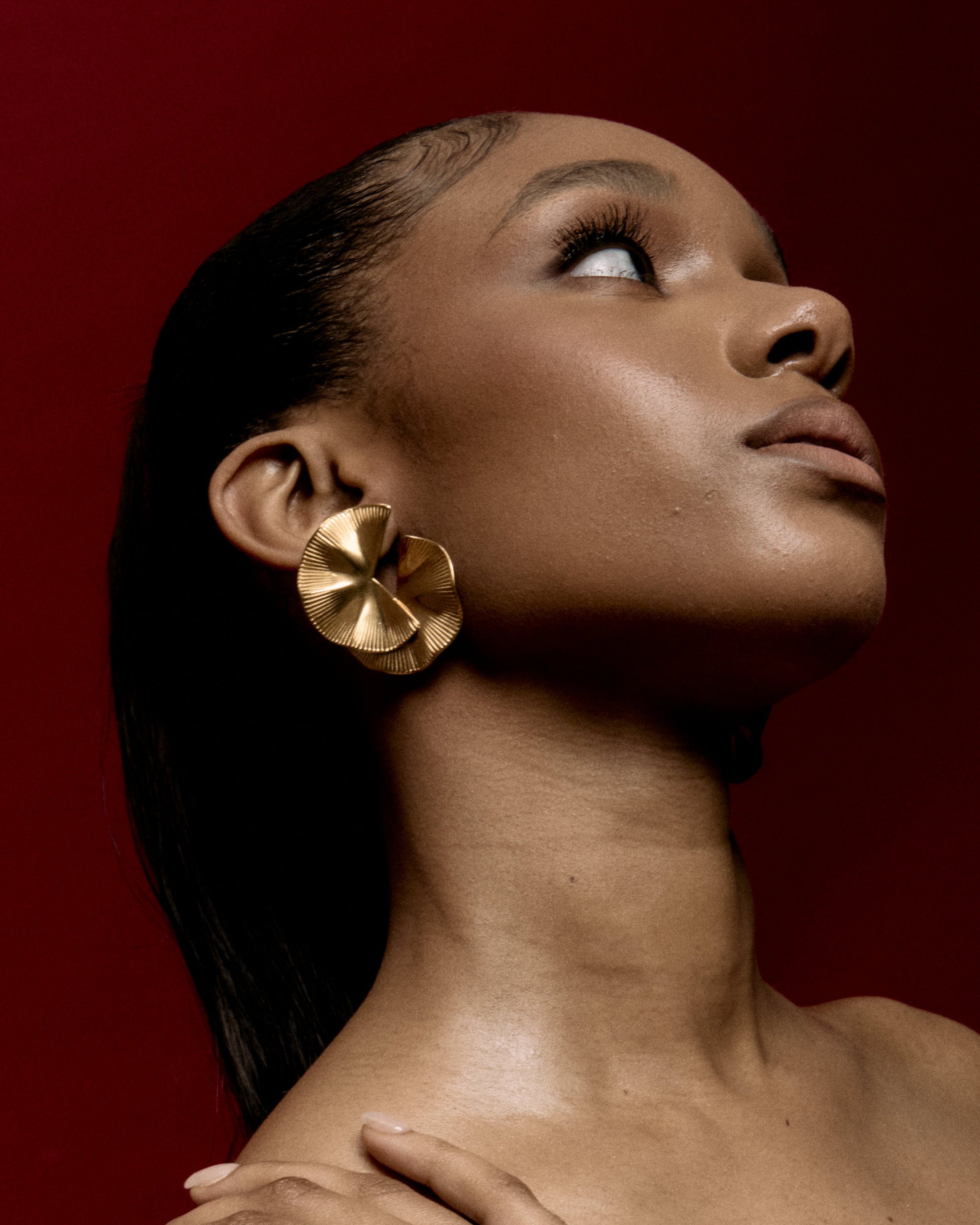 Close-up of a young woman with makeup, large gold earring, and shoulder-length dark hair against a dark red background.