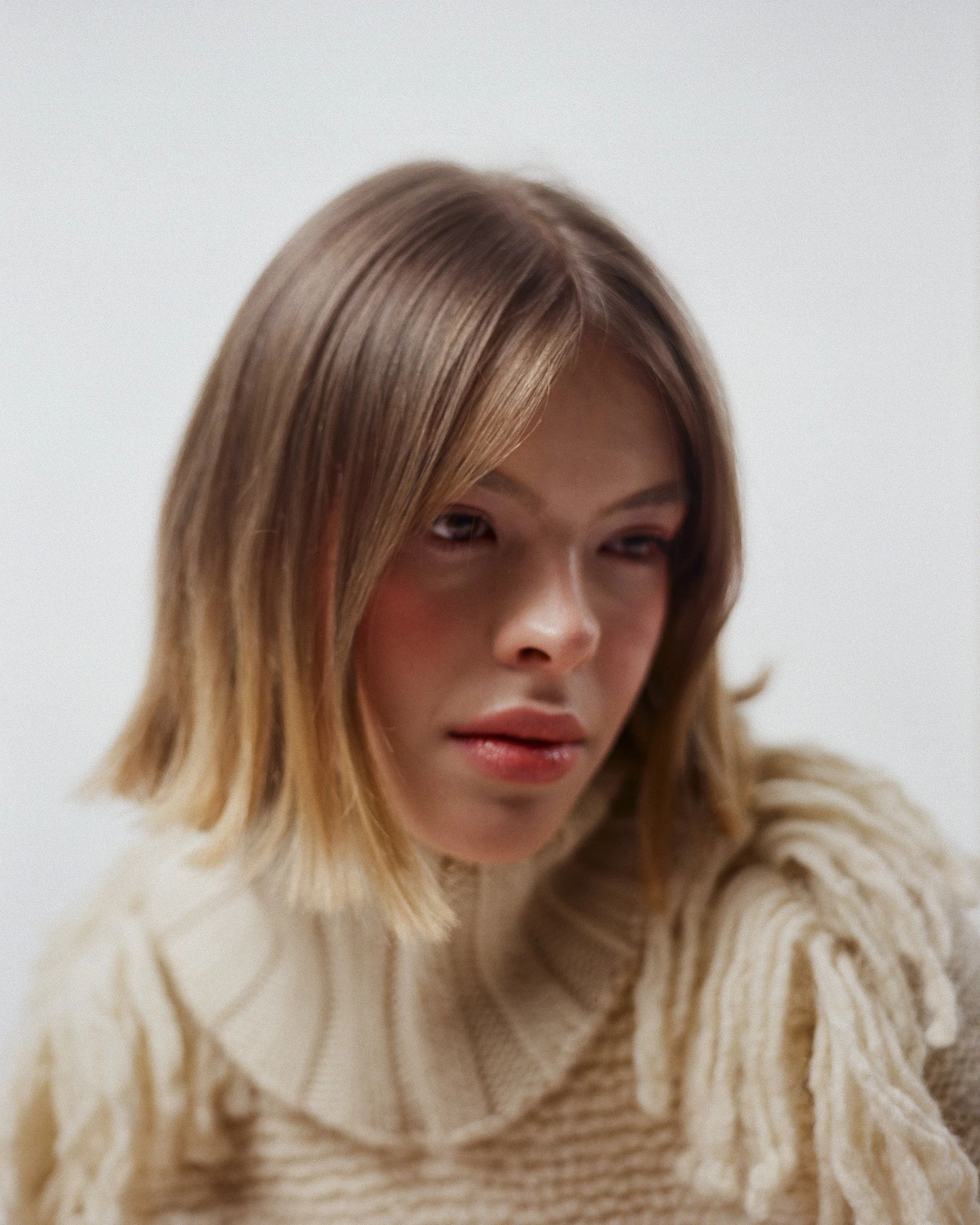 A young woman with light brown, shoulder-length hair, wearing a beige, cable-knit sweater with a turtleneck, looking downward with a neutral expression against a plain, light-colored background.