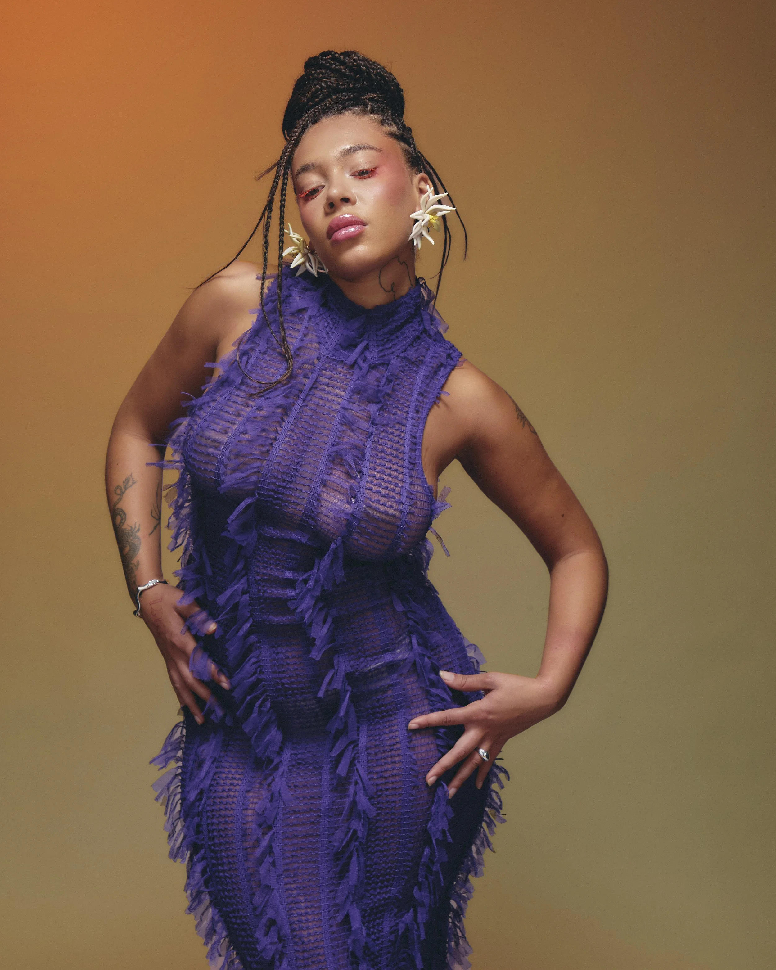 A woman with braided hair wearing a purple textured dress with fringes and large white floral earrings, posing against a gradient background.