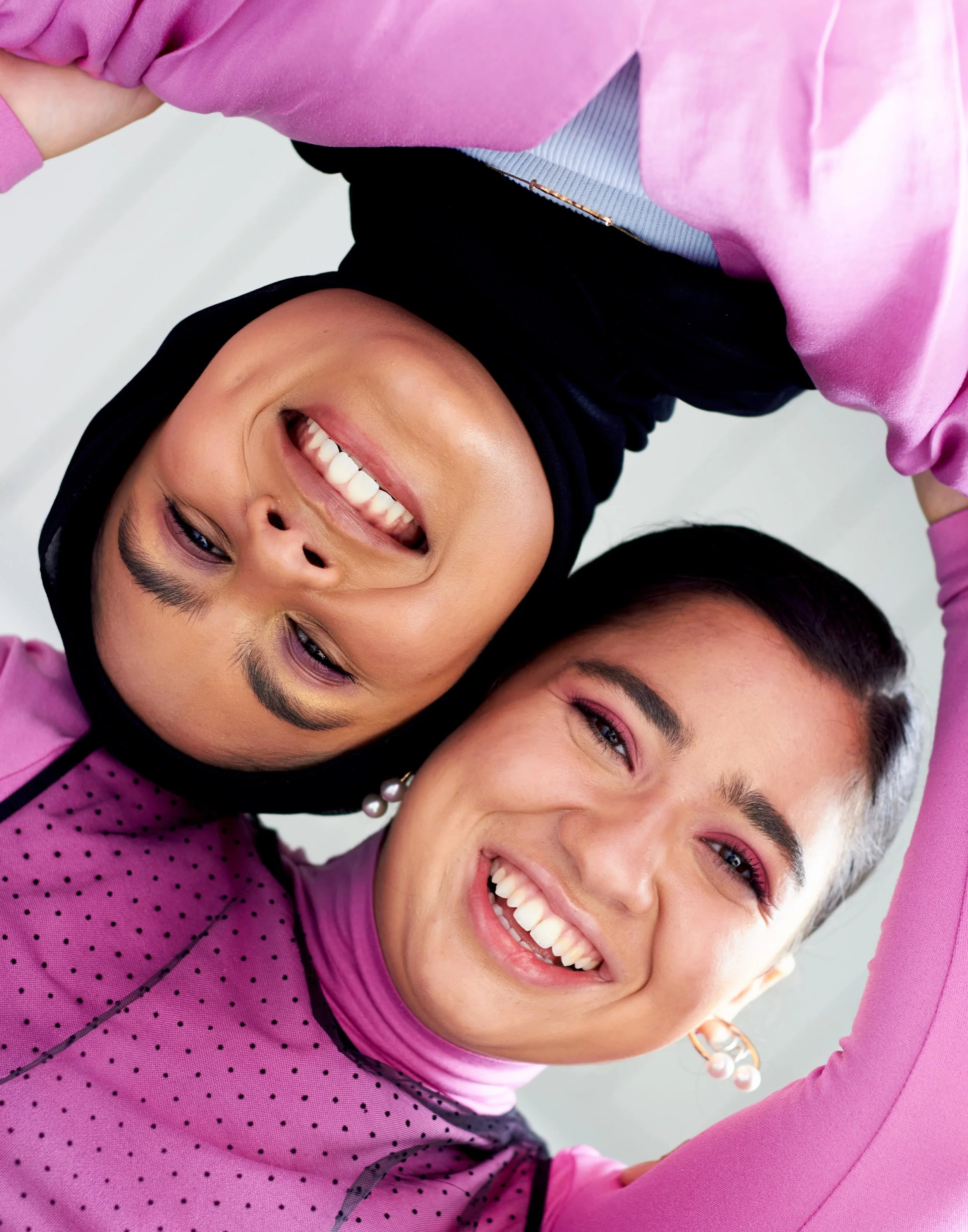 Two women smiling and taking a selfie, with one wearing a black hijab and the other with short hair and earrings.