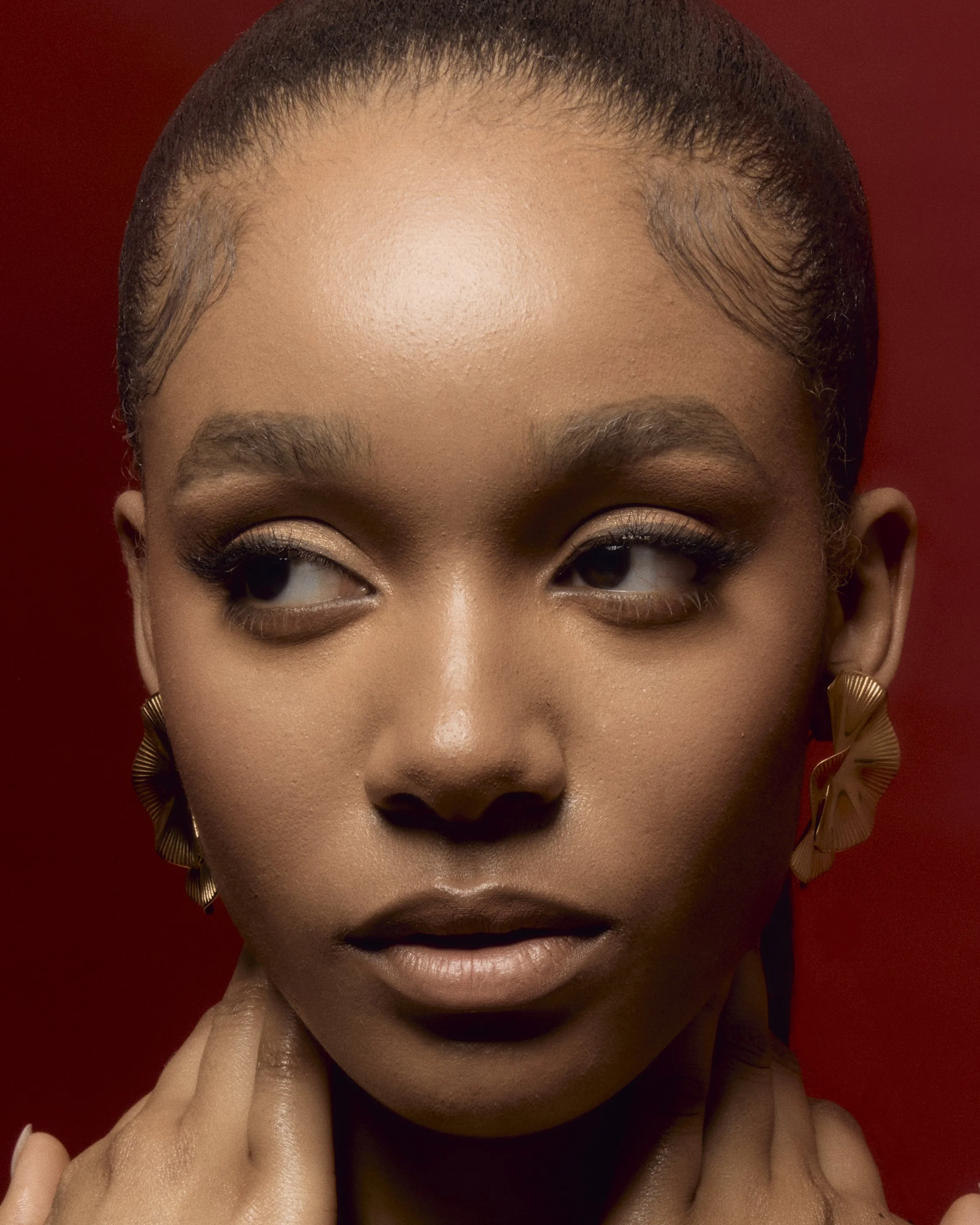 Close-up portrait of a woman with smooth skin and gold earrings, against a red background, with a contemplative expression.
