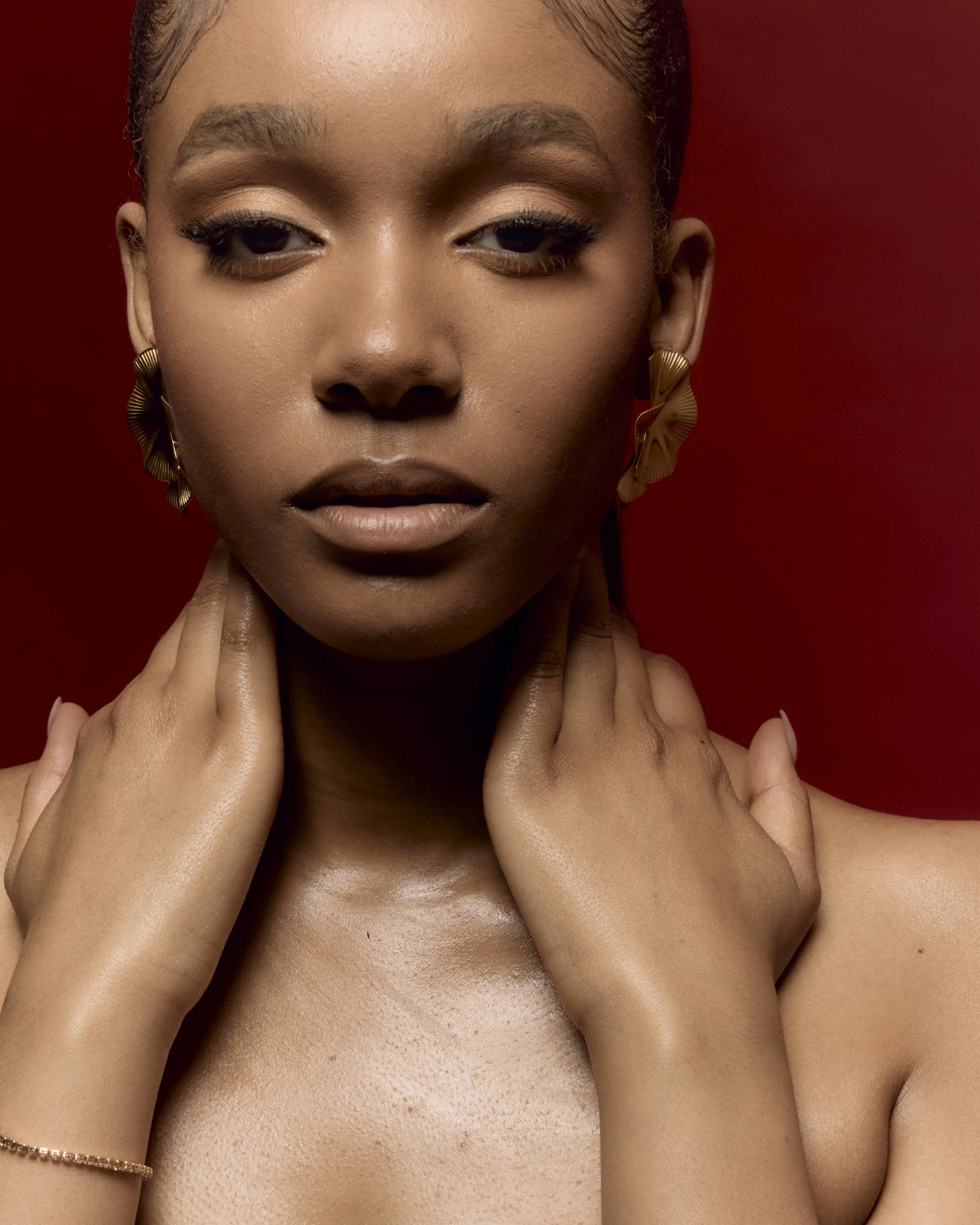 A close-up portrait of a woman with a dark complexion, gold earrings, and smooth skin against a red background.