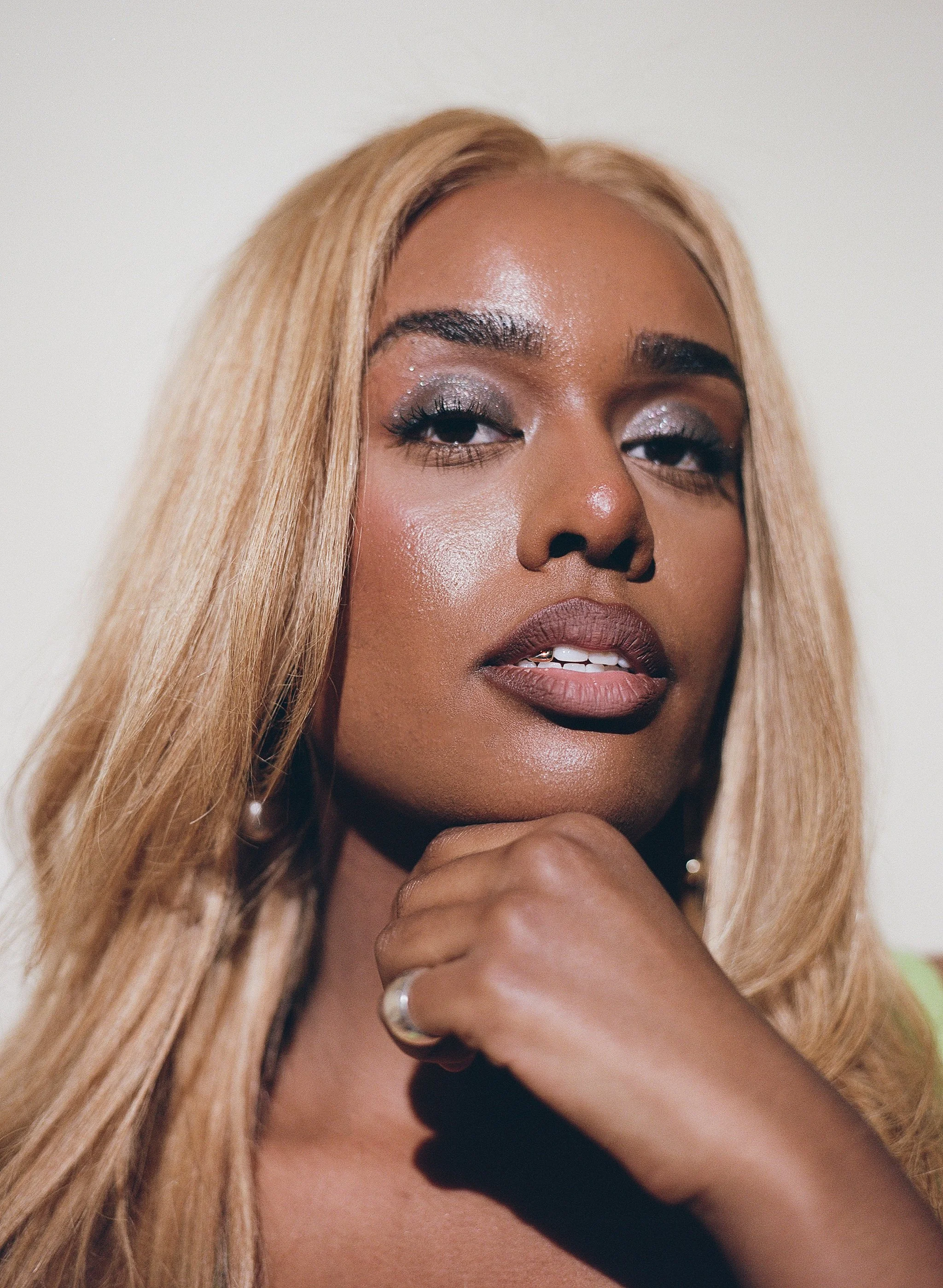 A close-up portrait of a woman with long blonde hair, wearing makeup with silver eyeshadow, dark lipstick, and a ring on her finger, resting her chin on her hand.