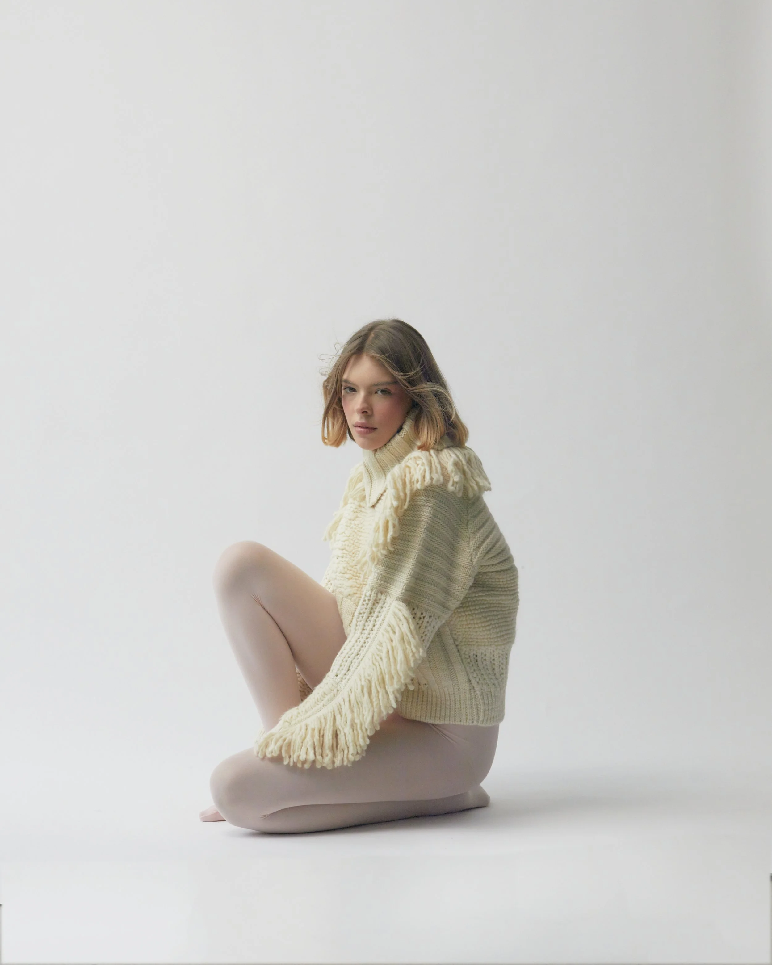 A young woman with shoulder-length light brown hair sitting on the floor against a plain white background. She is wearing a chunky cream-colored knit sweater and beige tights, looking at the camera with a serious expression.