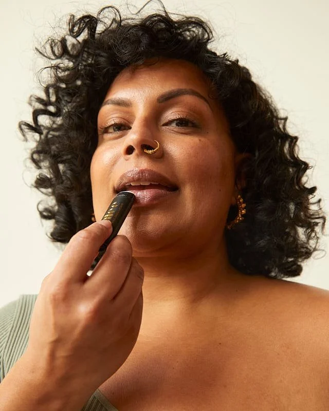 A woman with curly black hair wearing gold earrings and a nose ring, applying lipstick in front of a plain background.