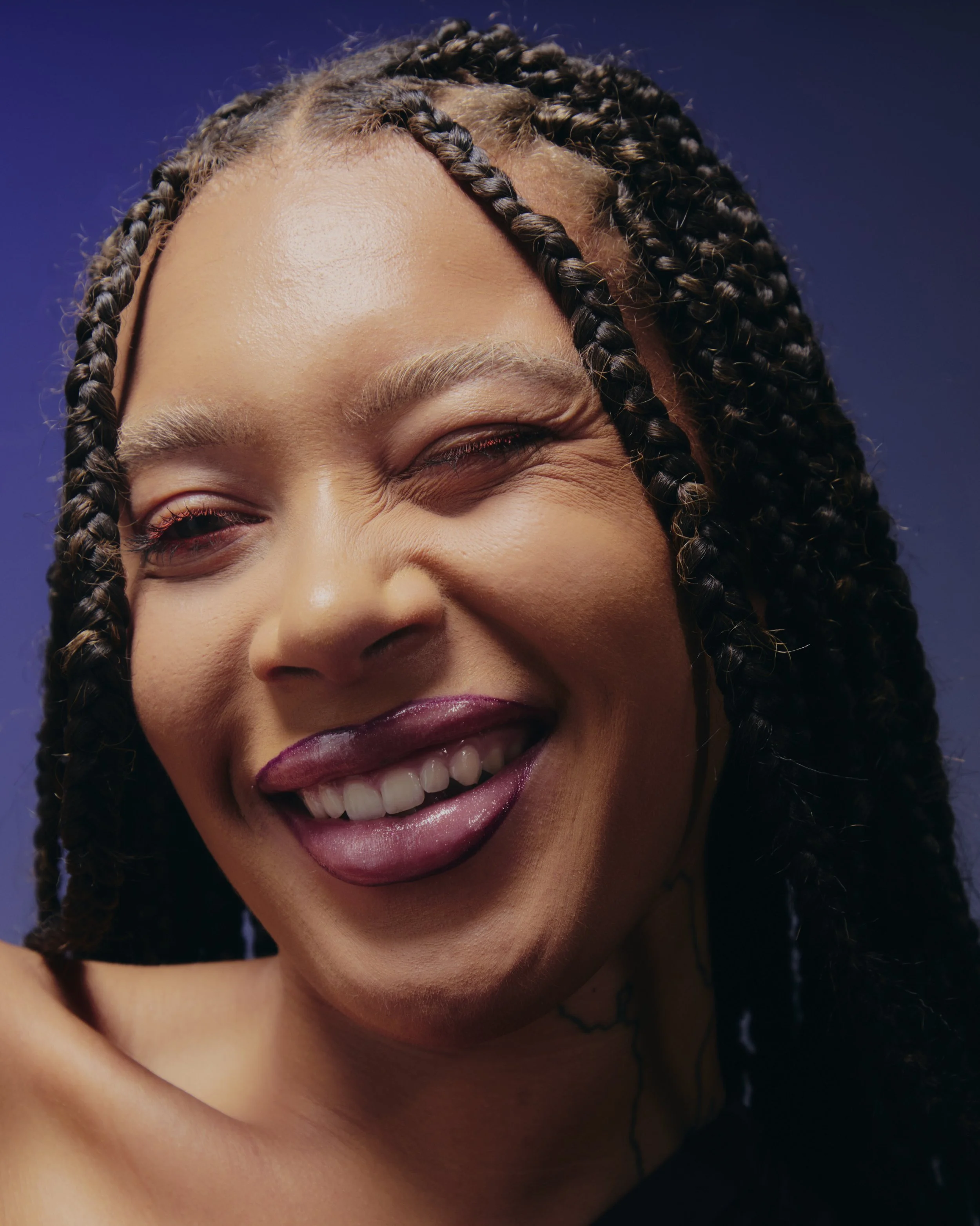 A close-up portrait of a smiling woman with long, braided hair, winking with her tongue slightly out, against a dark blue background.
