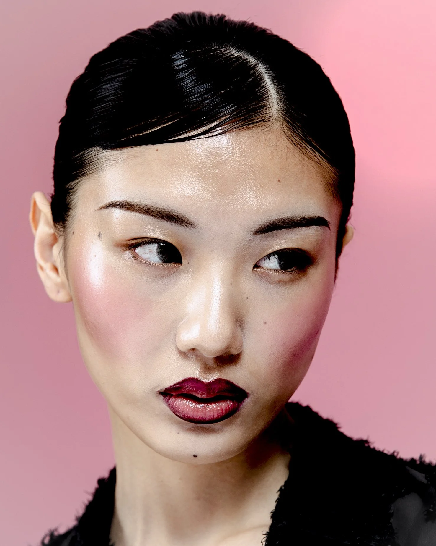 Close-up of a woman with black hair styled back, wearing makeup with dark lipstick, against a pink background.