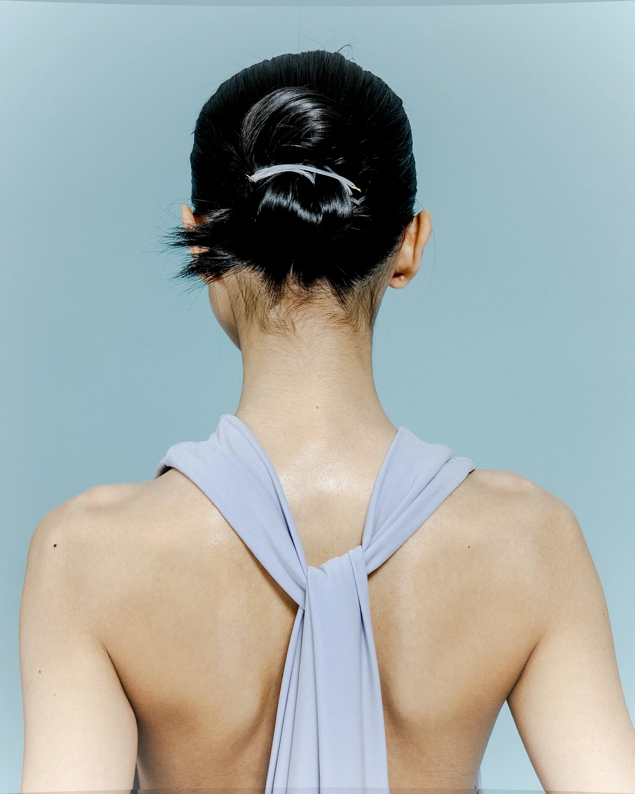 Back view of a woman with short black hair styled in an elegant updo, wearing a light gray dress with a bow at the back, against a blue background.