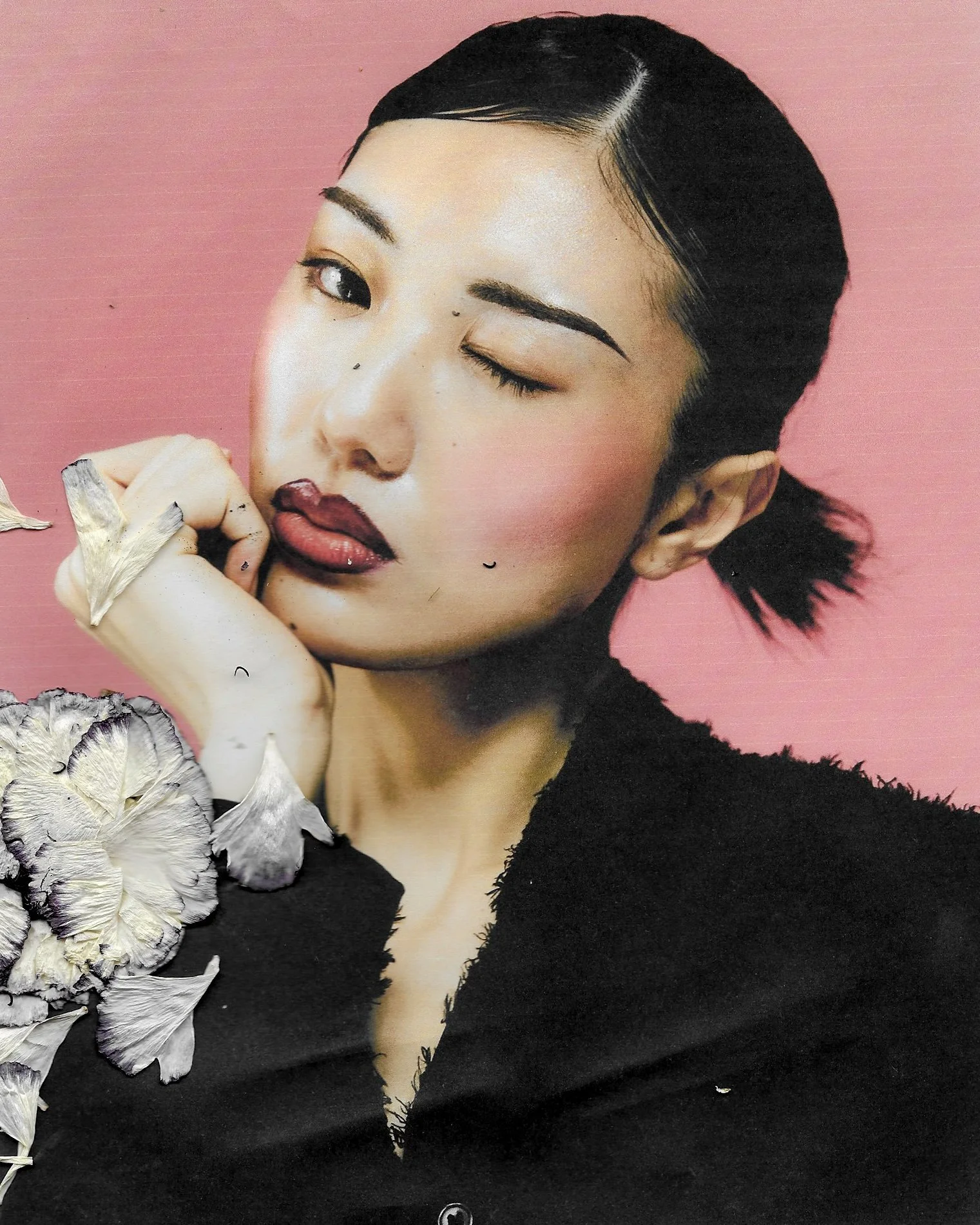 A woman with dark hair styled in a short bob, winking with one eye, wearing makeup, a black top, and holding dried flowers against a pink background.