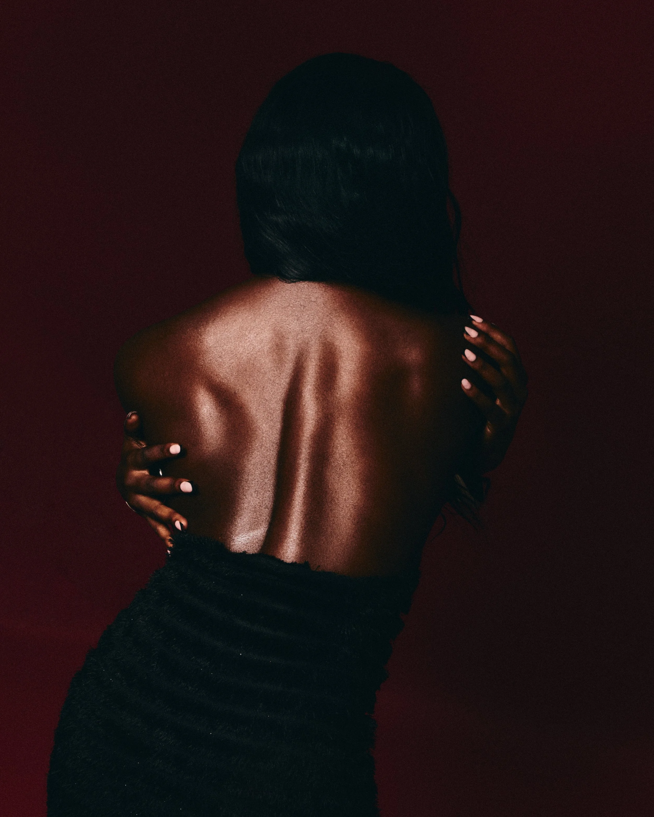 A woman with dark hair, seen from the back, hugging herself, with her arms crossed over her shoulders, against a dark red background.