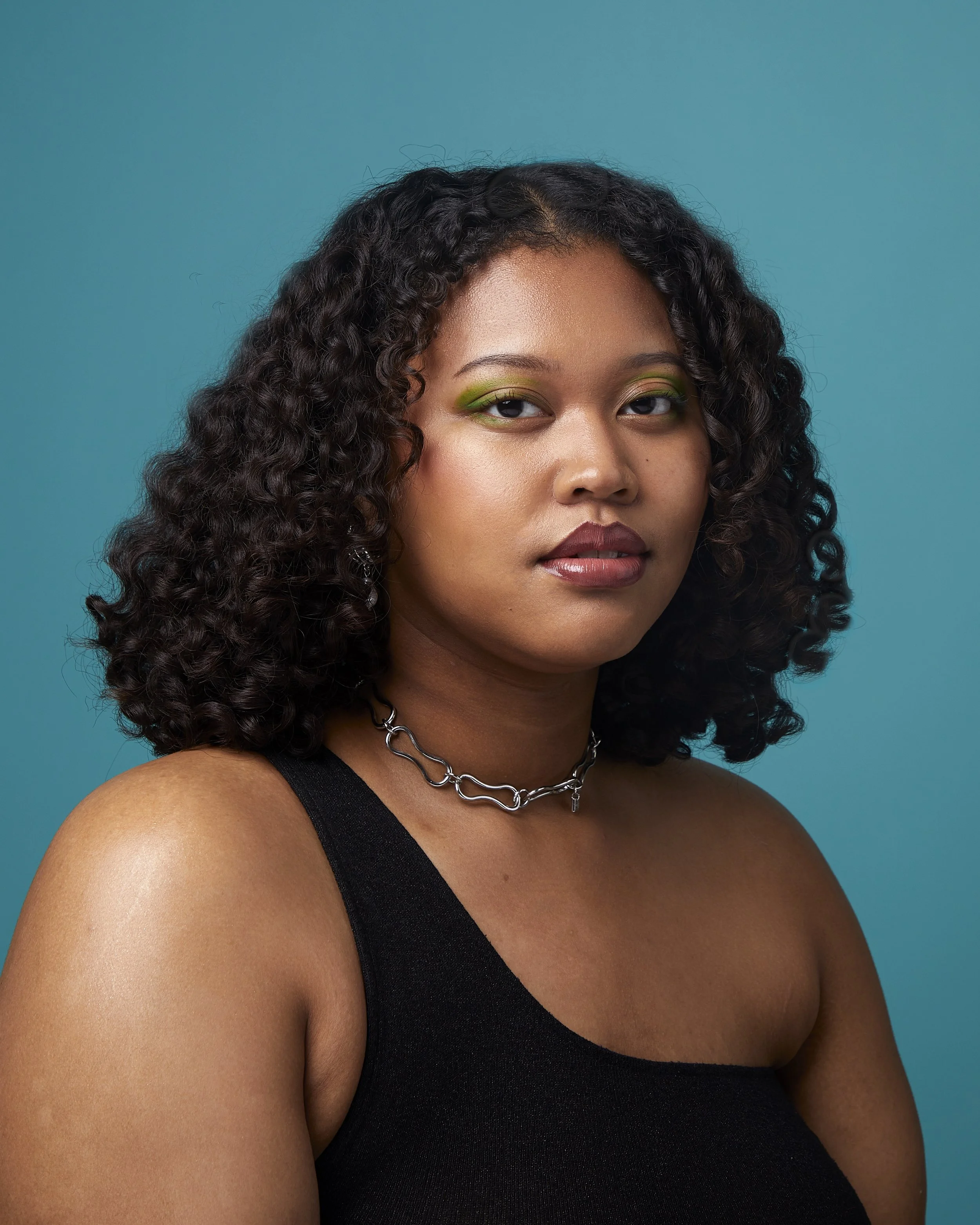 Portrait of a young woman with curly dark hair, wearing a black sleeveless top and silver chain necklace, against a teal background.