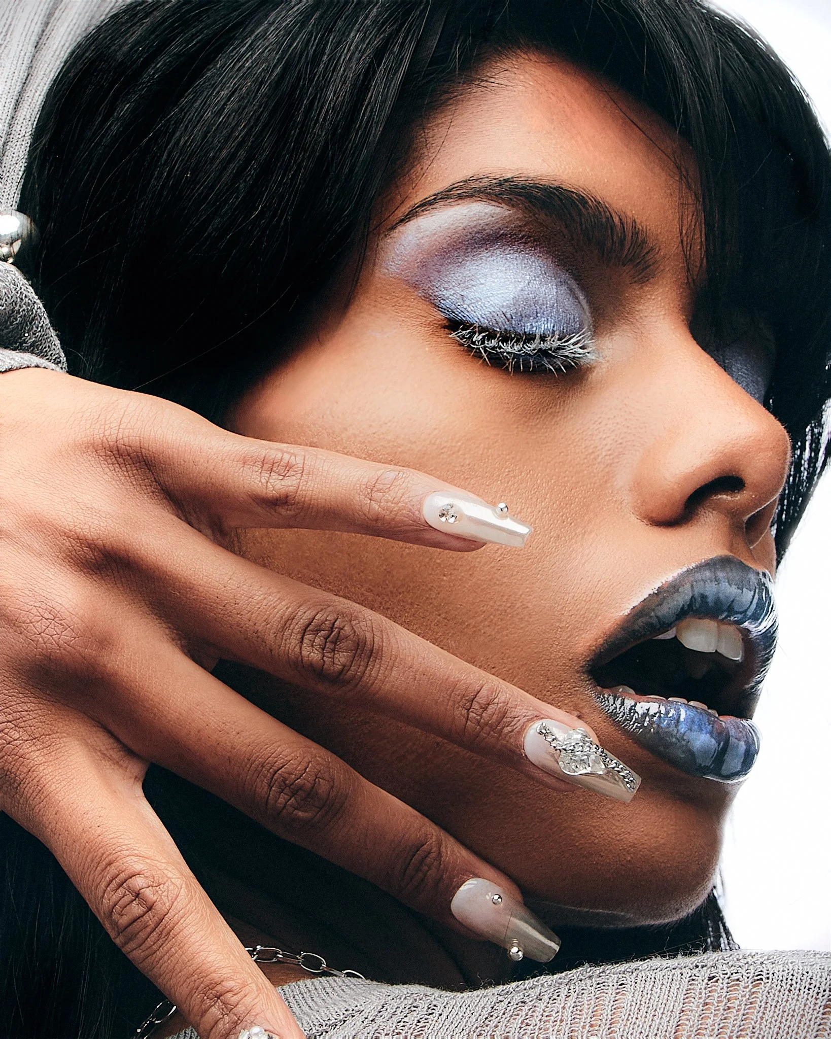 A woman with blue eyeshadow, black lipstick, and long nail art poses with her hand near her face.