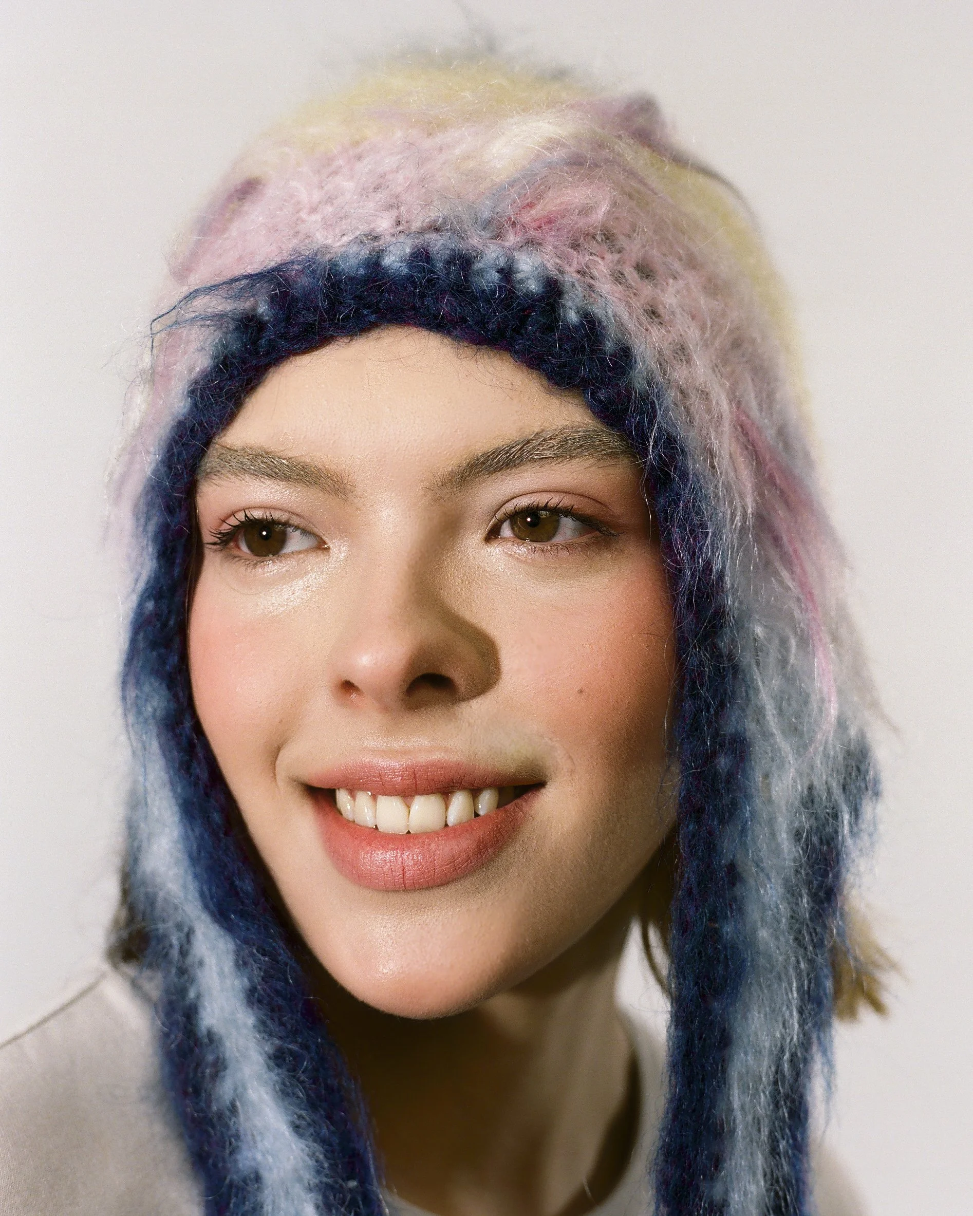 A young woman with multicolored, pastel pink, yellow, and blue dreadlocks, smiling with braces, against a plain white background.