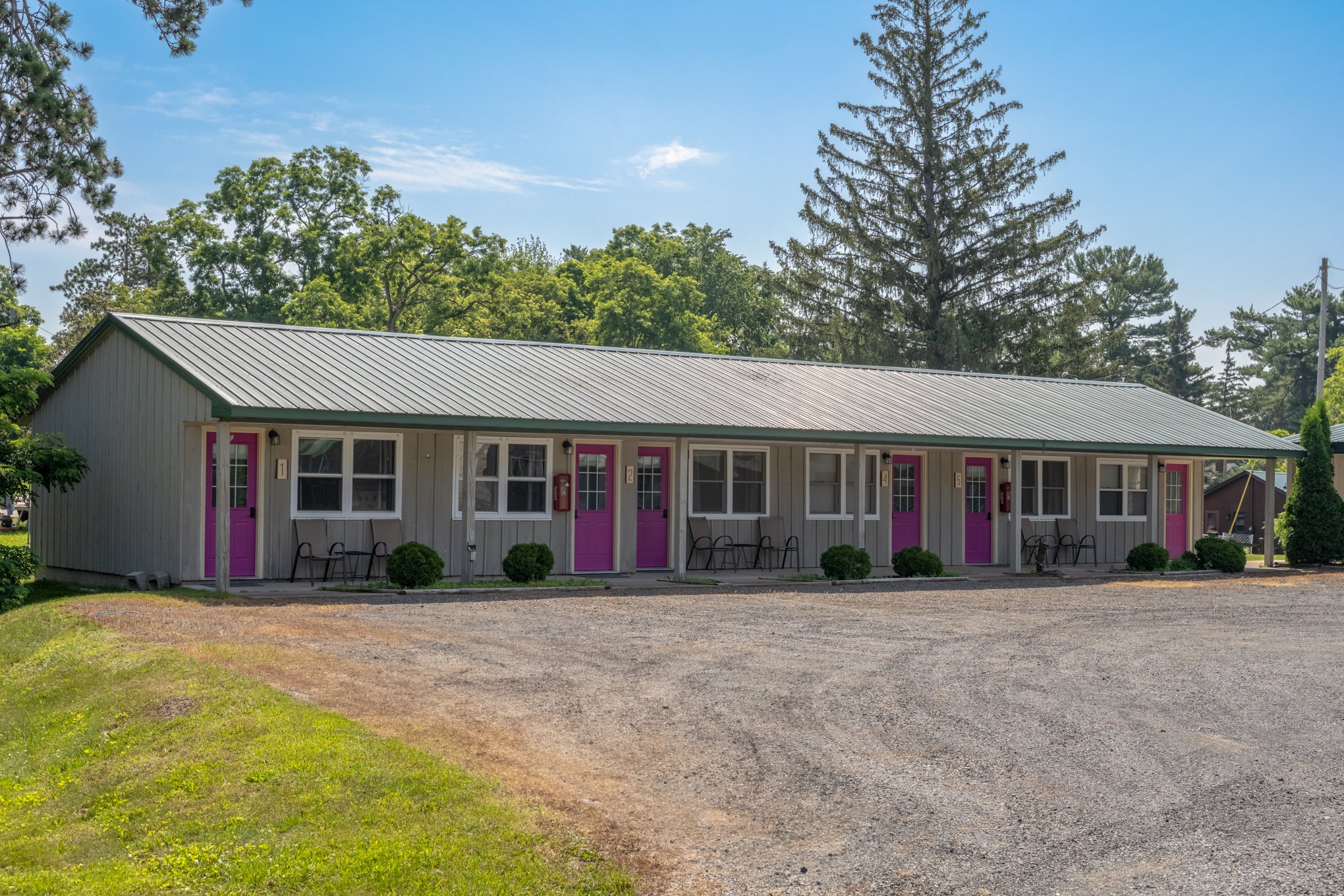 Single-story motel building with gray exterior and pink doors, surrounded by trees and a gravel parking area.