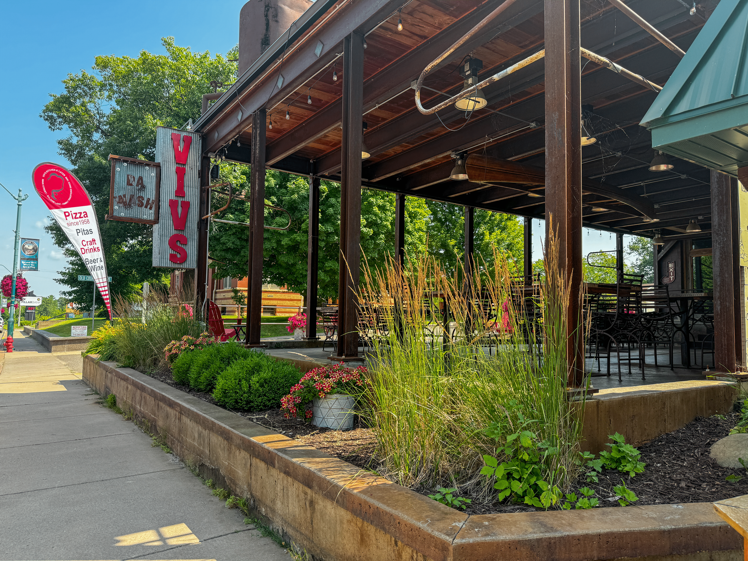 Outdoor restaurant patio with wooden beams and metal roof, featuring a vintage-style sign reading "VIVS" and "LA MASH." Sidewalk with plants and a red banner advertising pizza, craft drinks, beer, and wine. Sunny day with green trees in the backgroun