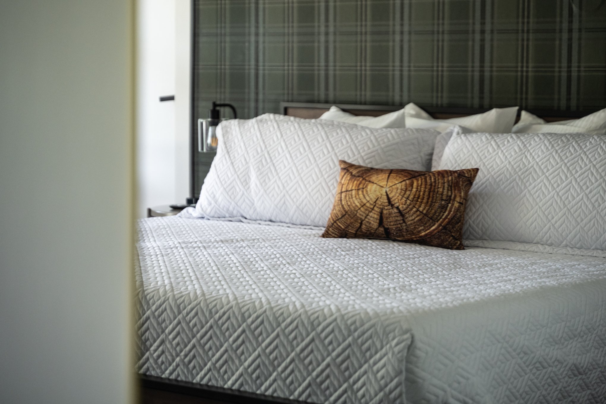 Bedroom with plaid wallpaper, white quilted bedding, and a decorative pillow resembling a tree log cross-section.