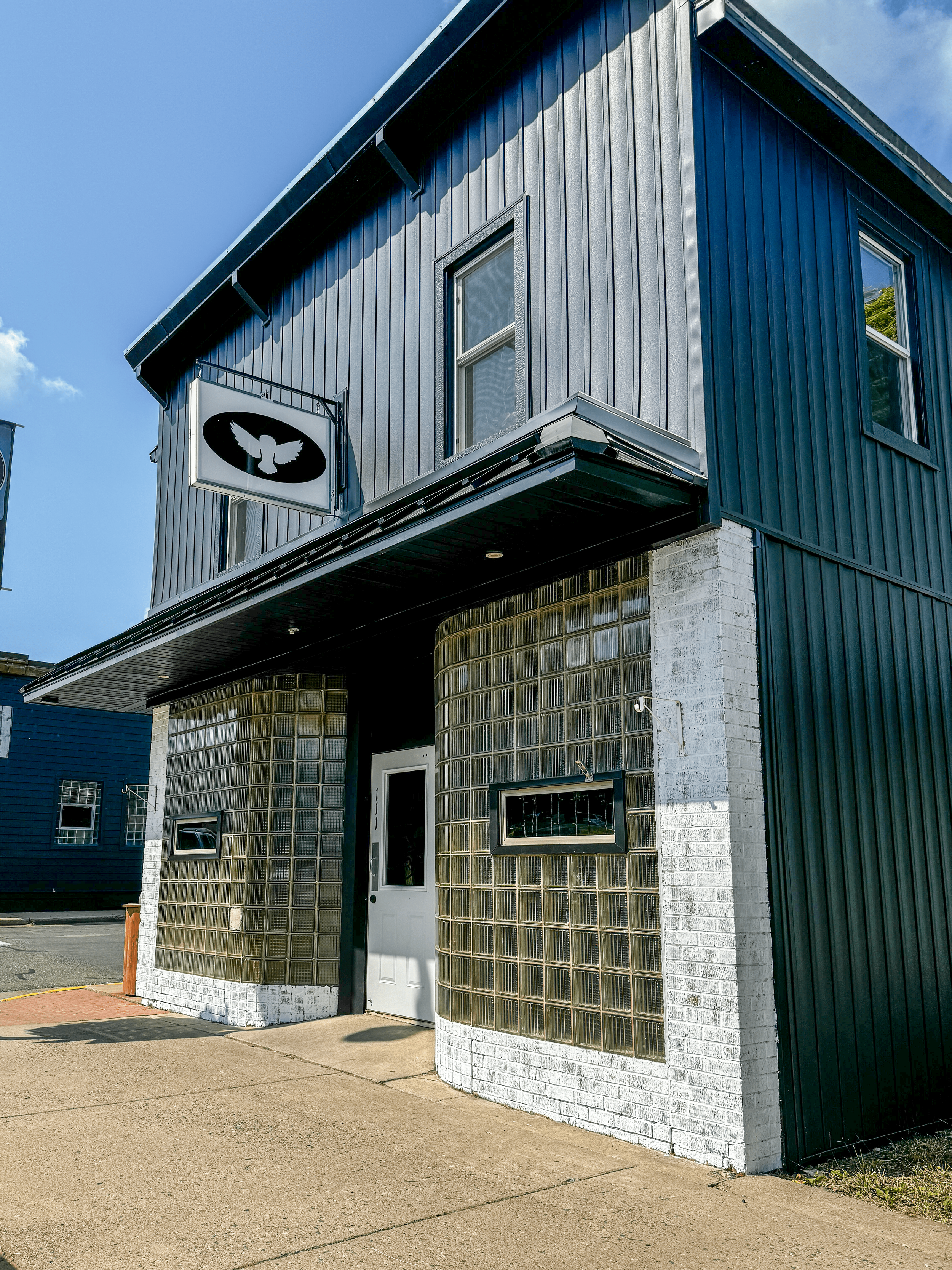 Two-story blue building with metal siding, glass block windows, and a white door. A sign with a stylized logo hangs above the door. Sidewalk and street visible.