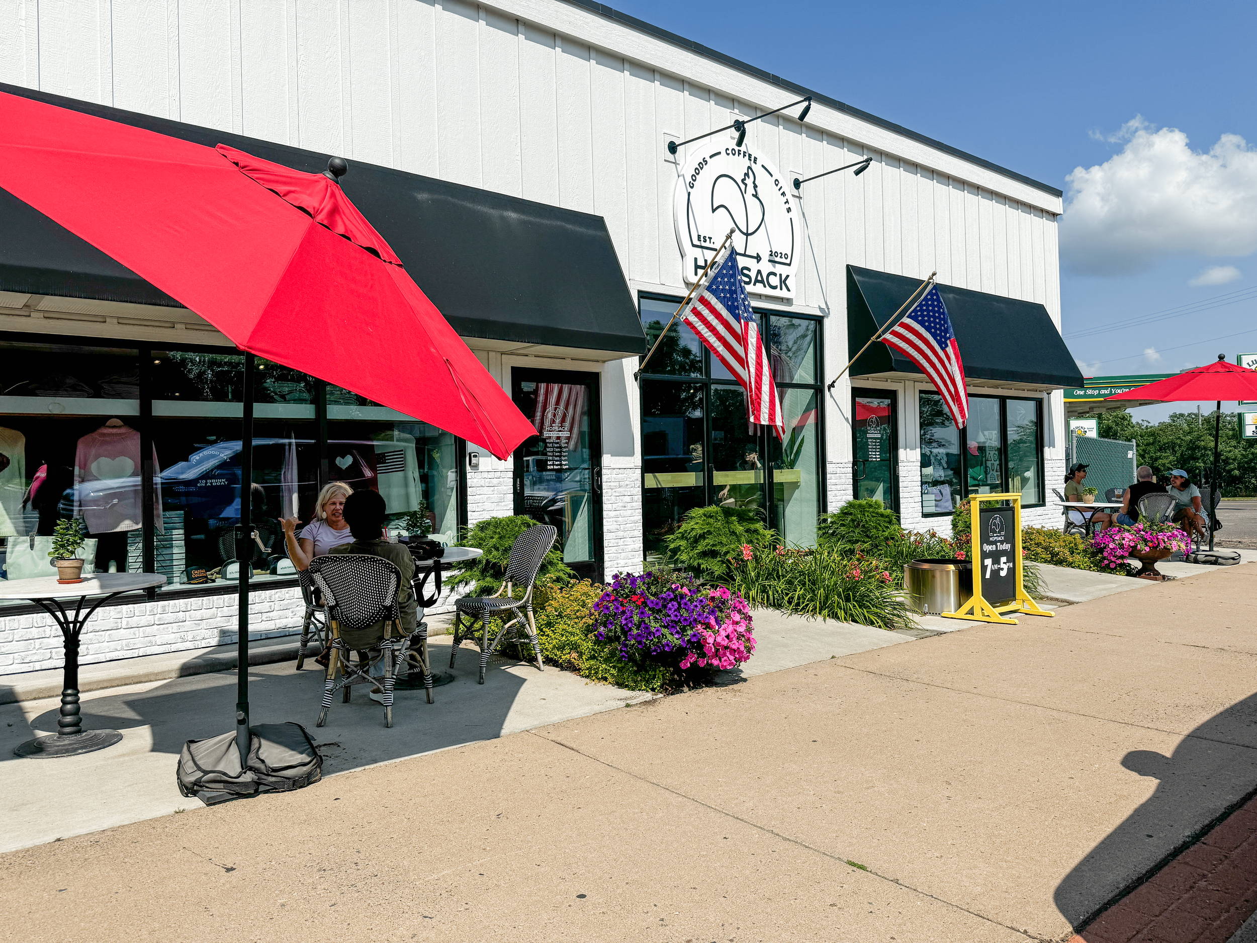 Outdoor cafe with red umbrellas and American flags outside a white building. People sitting at tables with chairs under umbrellas. Flower pots decorate the sidewalk.