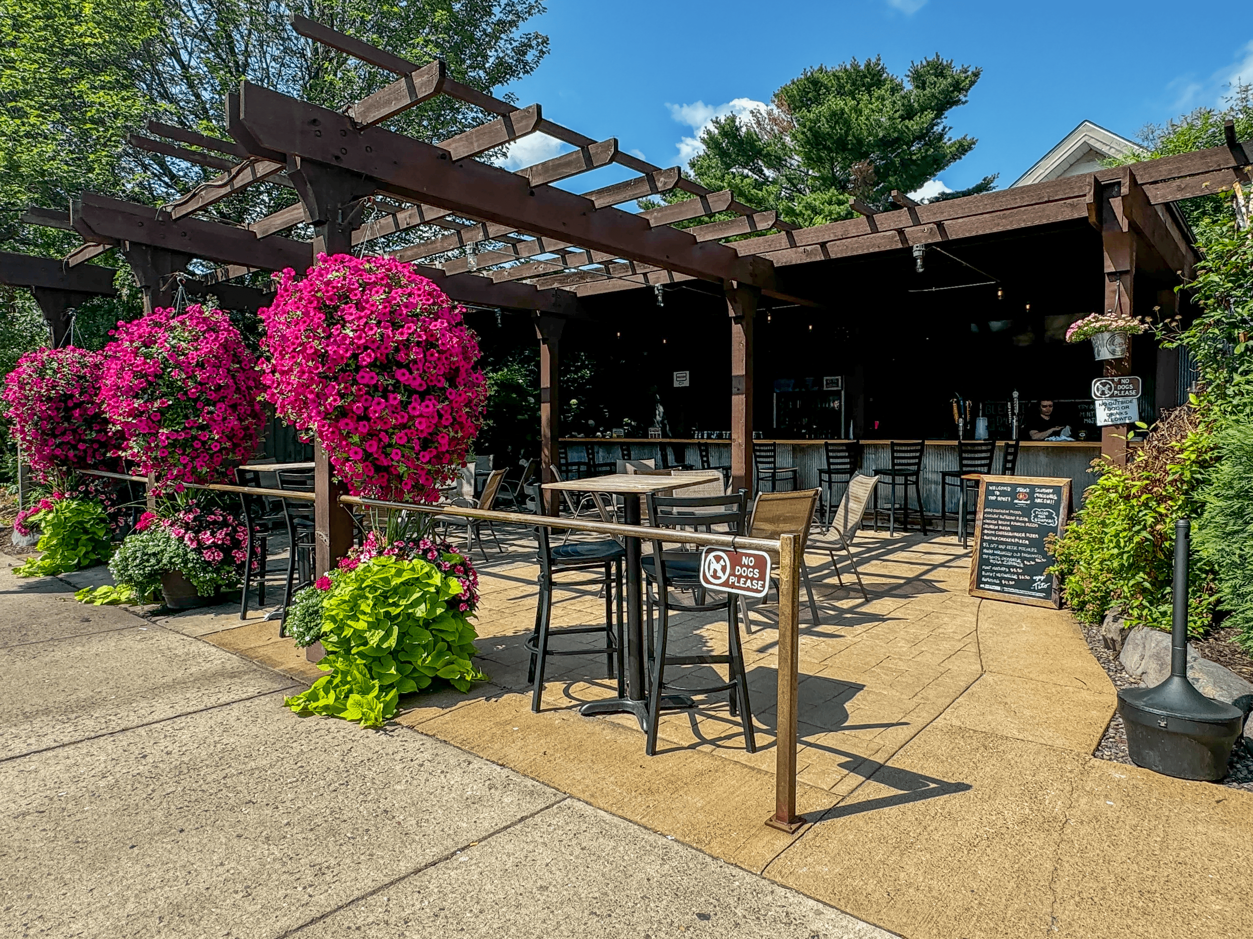 Outdoor restaurant patio with wooden pergola, vibrant pink flowering plants, tables and chairs, bar area, and a menu board.