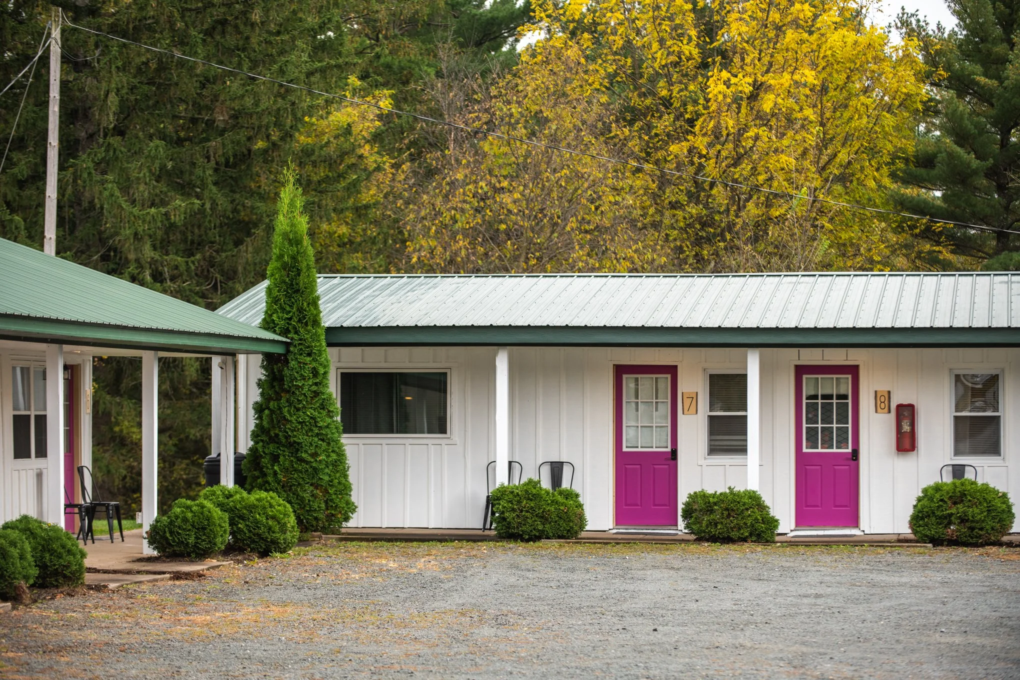 Exterior view of the motel, each featuring a bright pink door, chairs on the porch, and a green metal roof. The surrounding area has trees and a gravel parking lot.