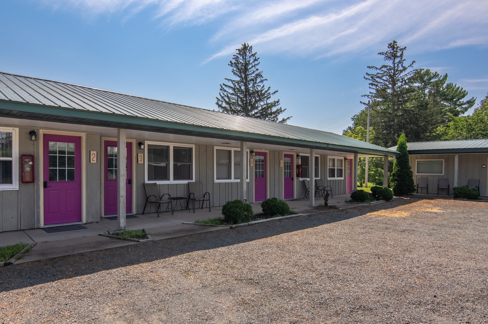 Exterior view of a motel with four units, each featuring a bright pink door, chairs on the porch, and a green metal roof. The surrounding area has trees and a gravel parking lot.