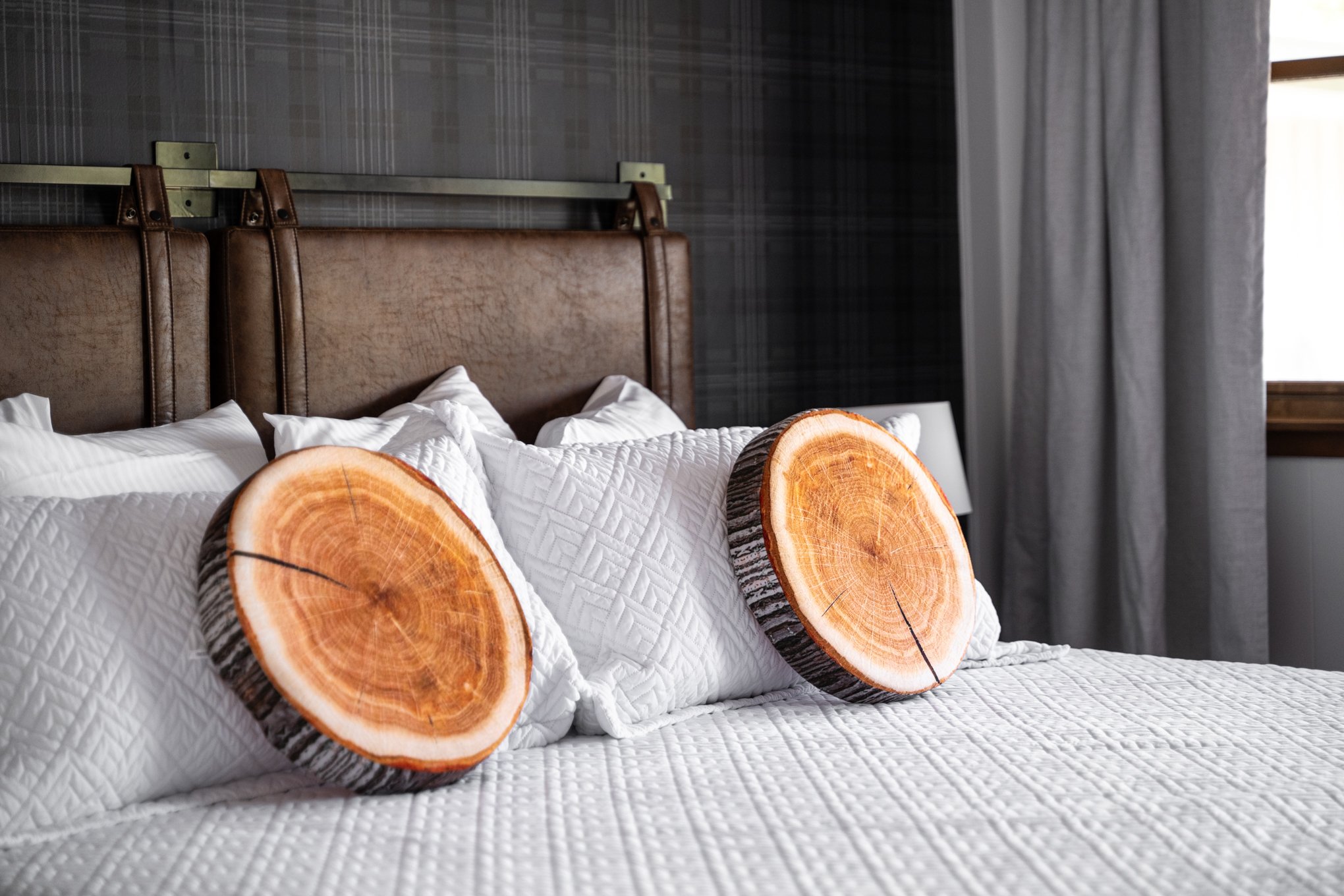 Bedroom with bed featuring white quilted bedding and pillows, log slice-shaped cushions, leather headboard, and dark plaid wallpaper.