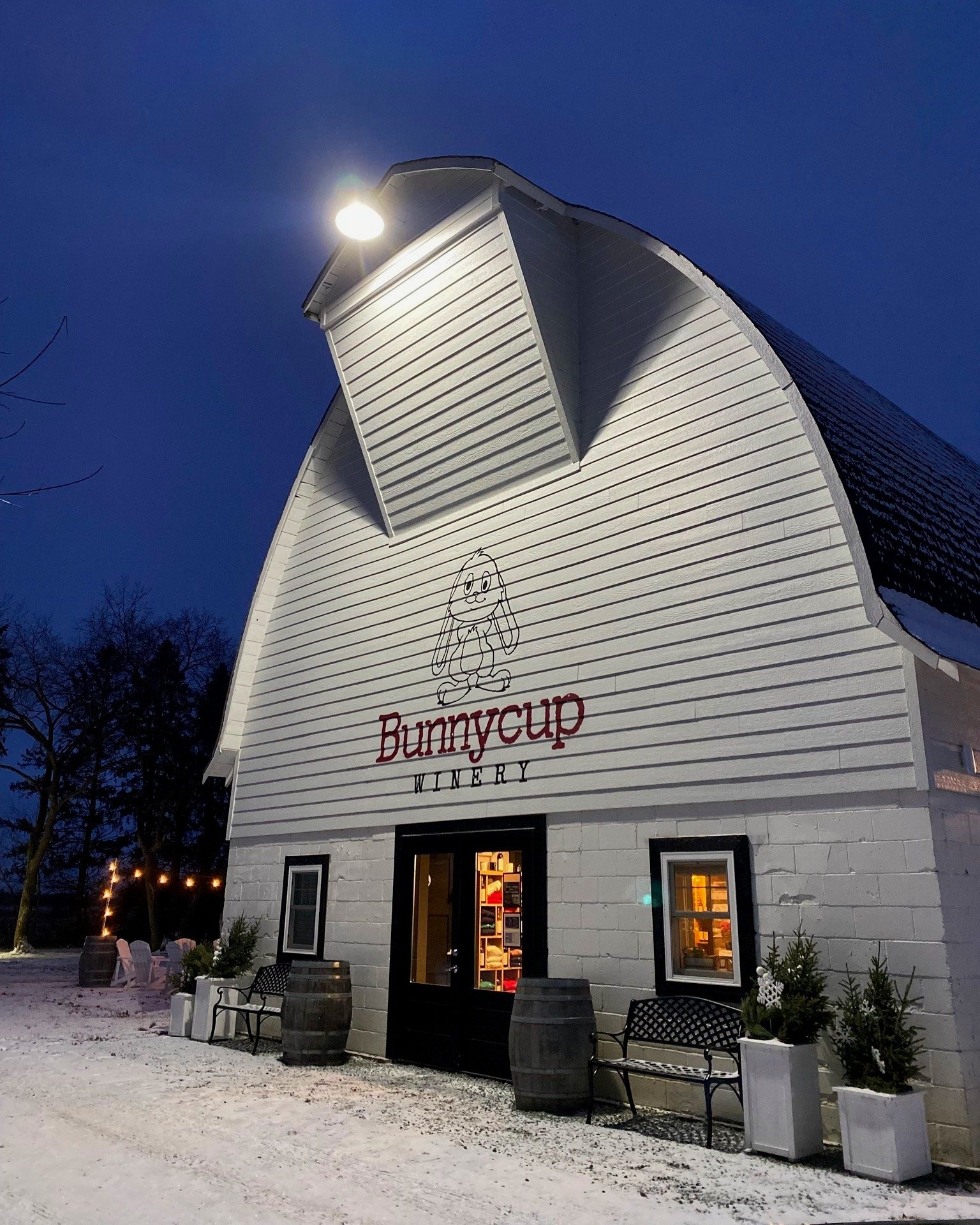 Bunnycup Winery building at night, with snow, benches, barrels, and string lights.