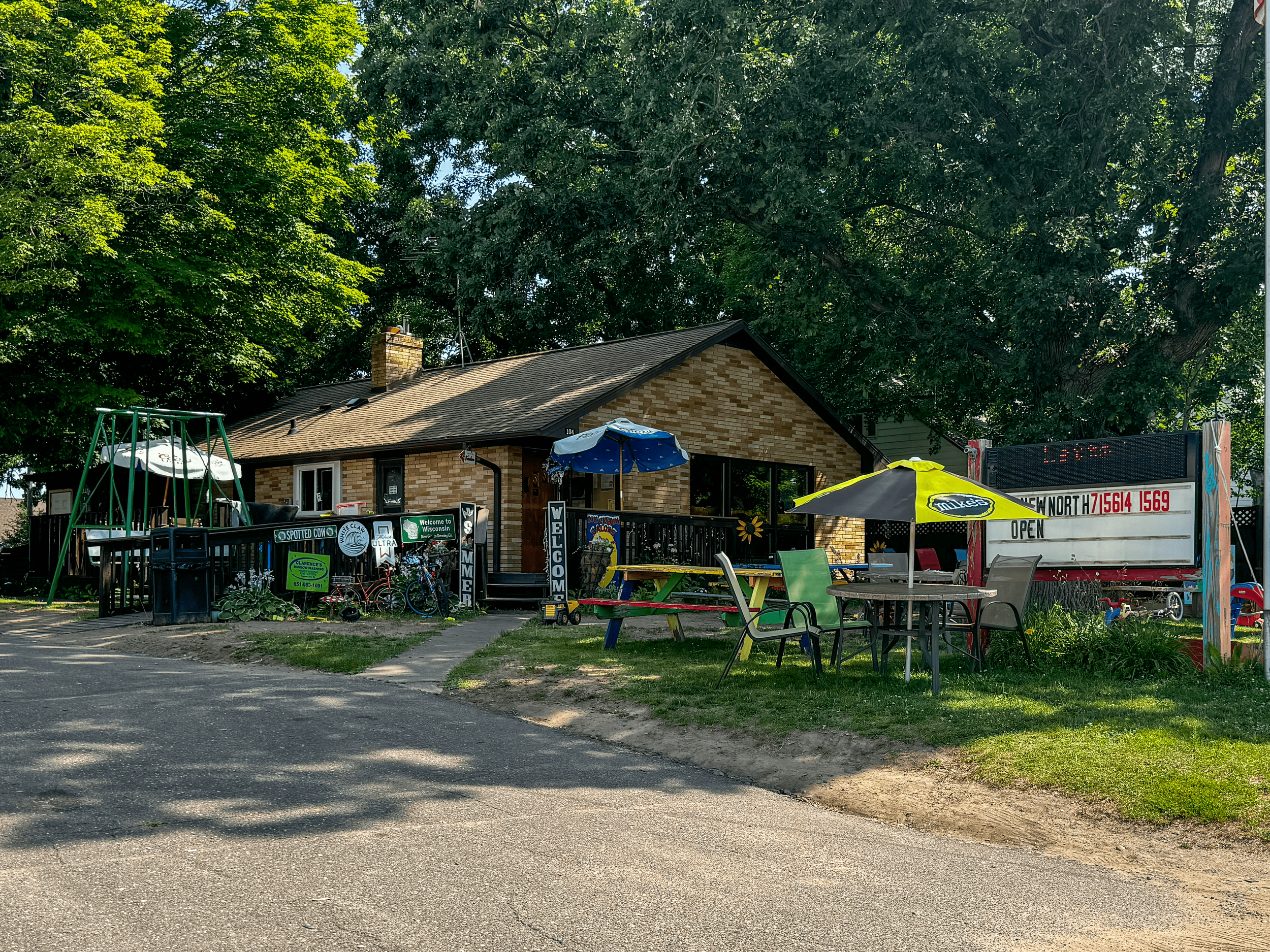Outdoor view of a small brick building surrounded by trees, with a deck and patio seating. Several chairs and tables with umbrellas are visible. A sign displays "Open" along with numbers. Playground equipment and various signs are also present.