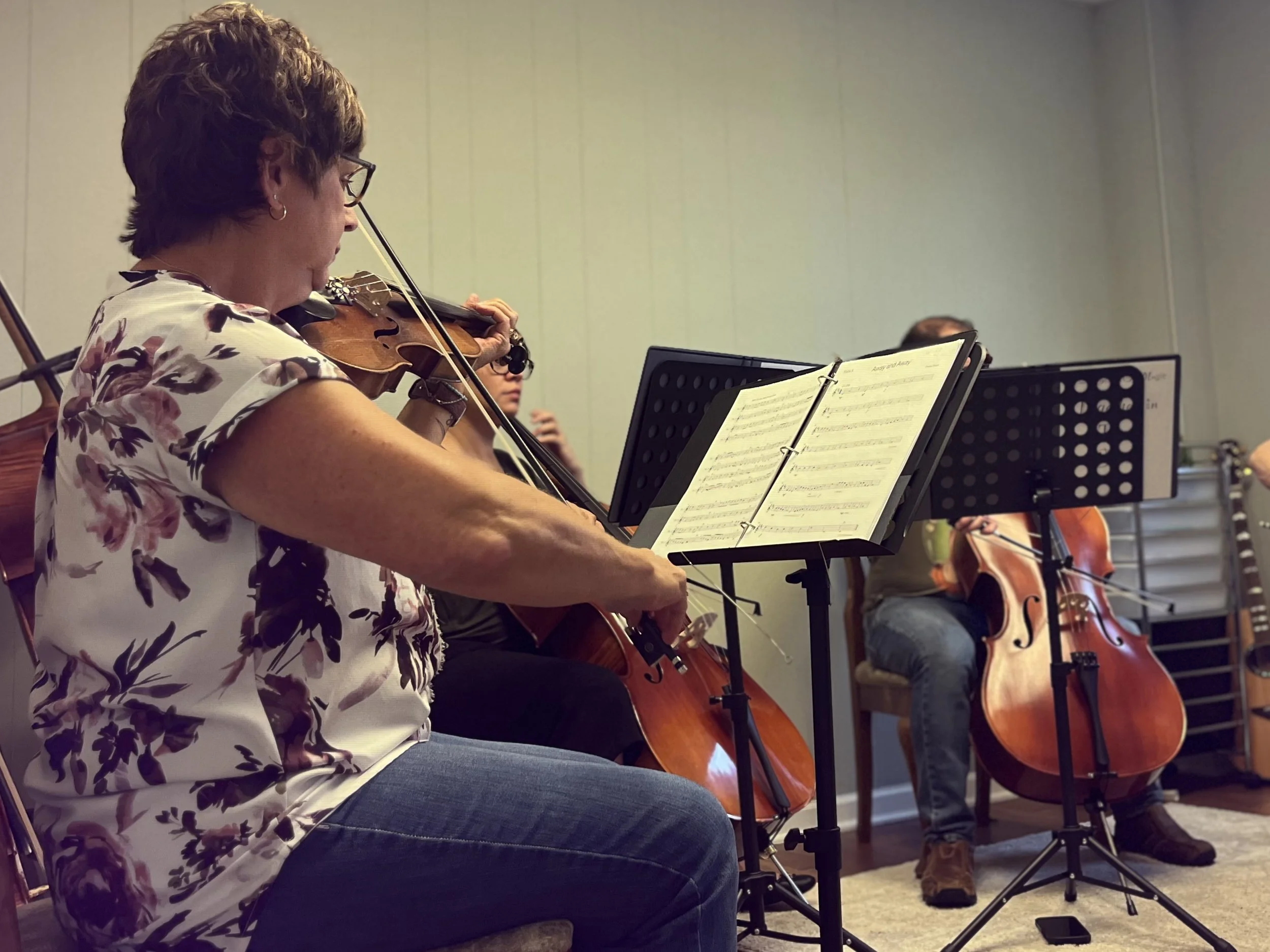 Three musicians playing string instruments during a practice session or performance, with sheet music on stands in front of them.