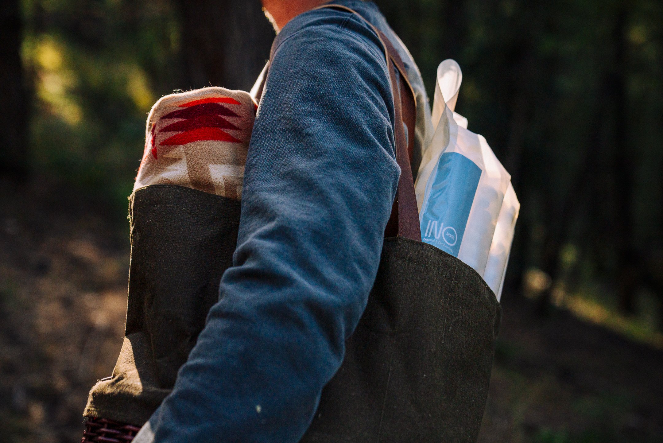 A person in a blue long-sleeve shirt carrying a black tote bag with a rolled-up blanket inside and a tote bag with a water bottle in an outdoor wooded area during daylight.