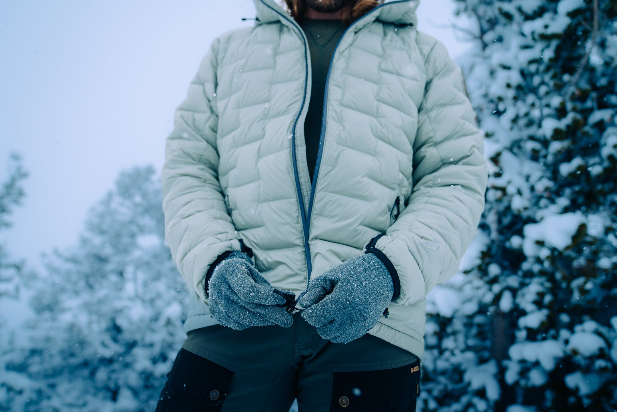 A person wearing a light-colored puffy jacket, gray gloves, and dark pants standing outdoors in a snowy environment with snow-covered trees in the background.