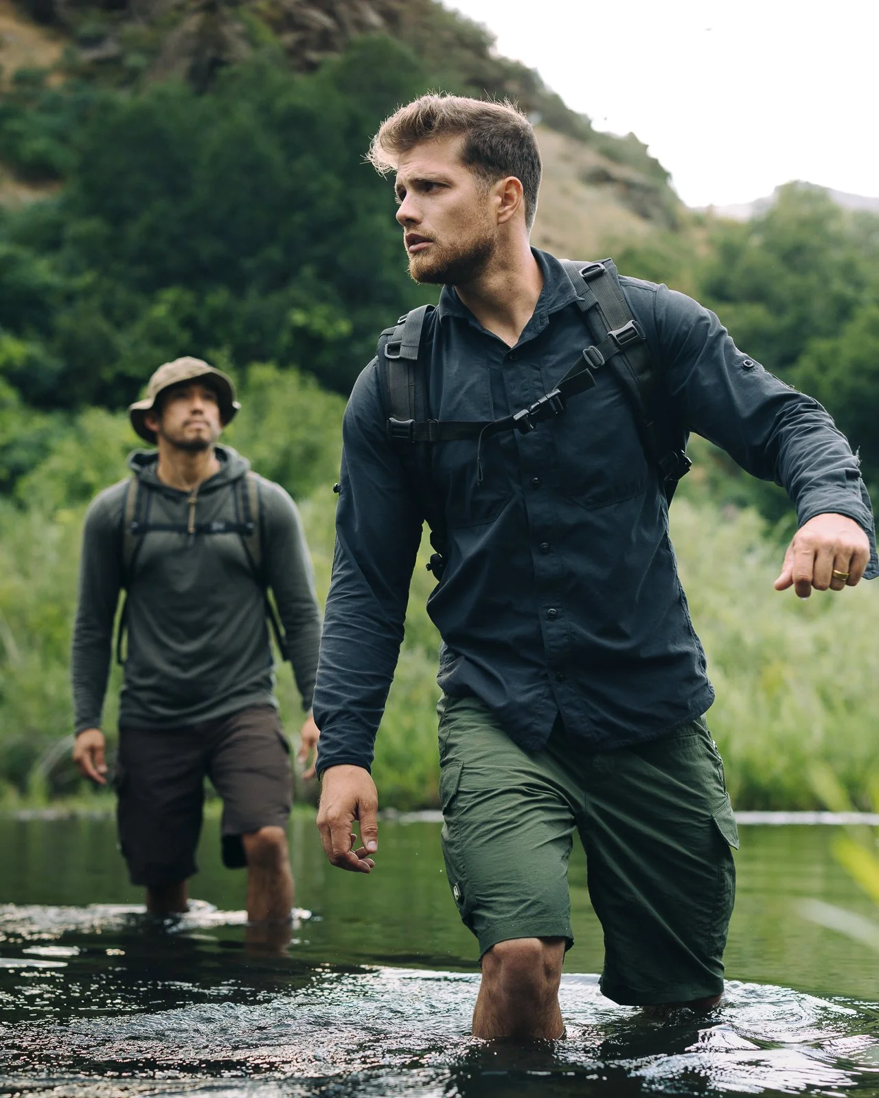 Two men hiking through a shallow river in a green, mountainous forest.