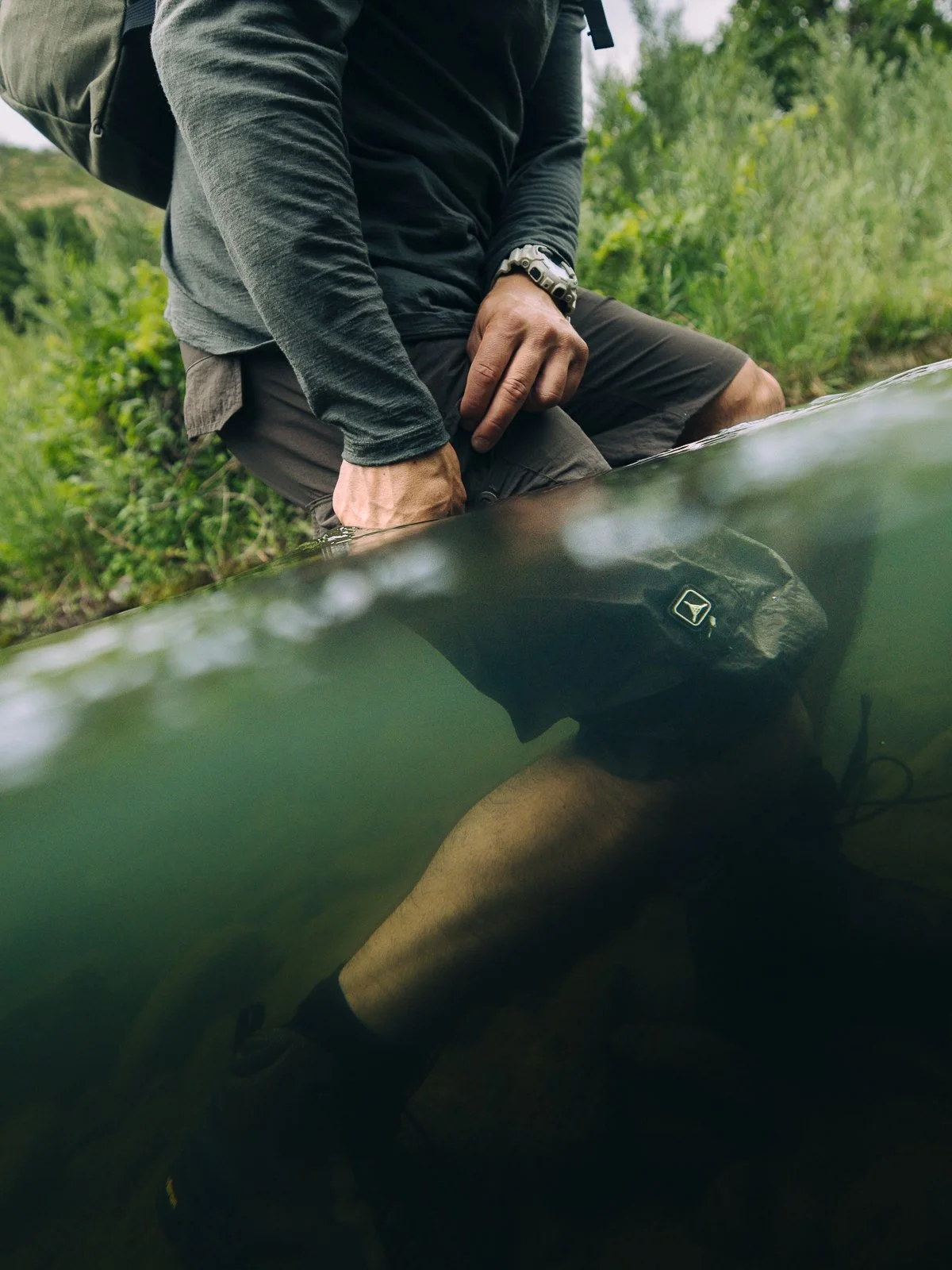 A person kneeling by the water's edge in a natural outdoor setting, with part of their body underwater visible below the surface. Amphibious cloth made for adventure. Product shot of shorts.