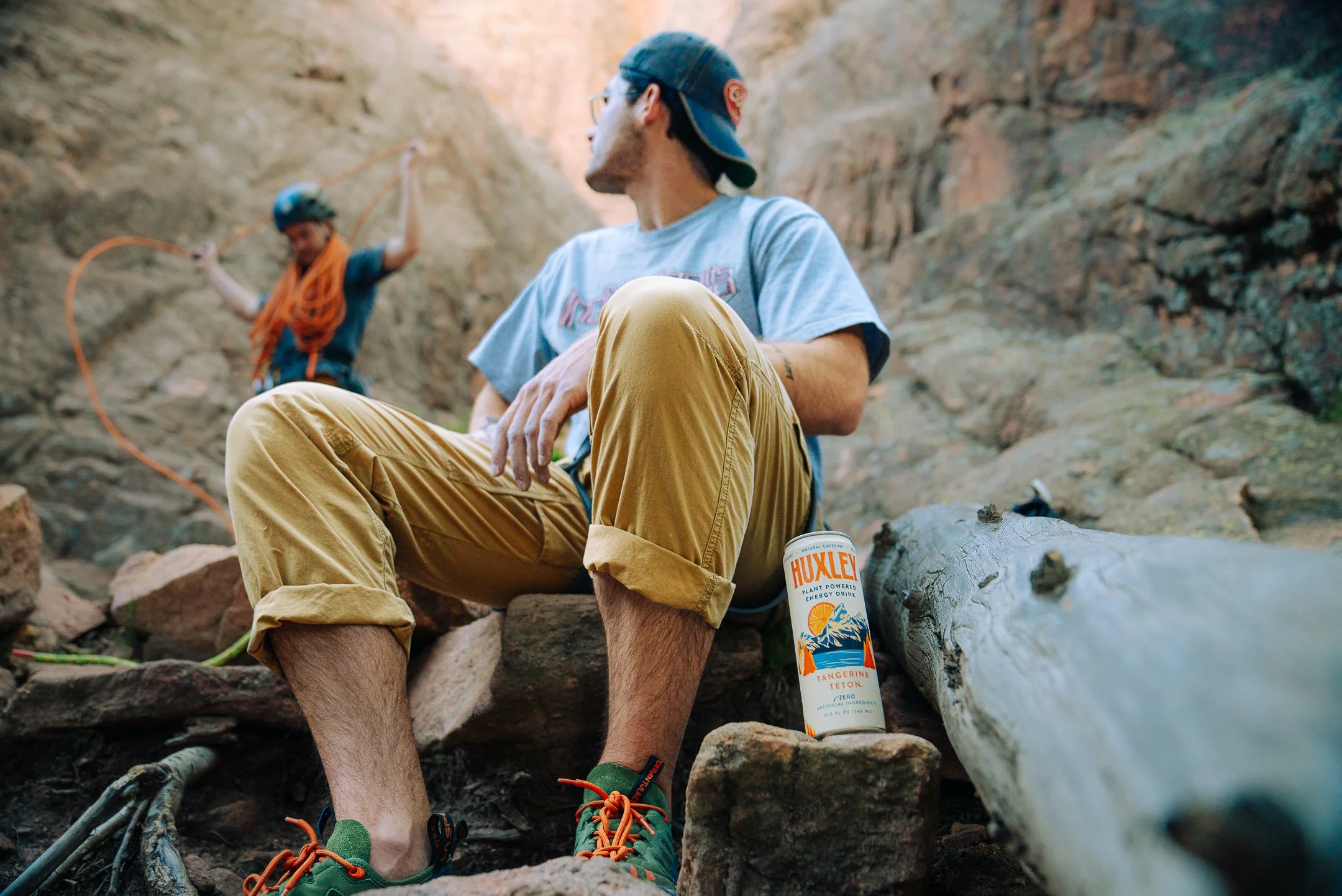 Two hikers resting in a rocky canyon. The man in the foreground sits on a rock, wearing a blue cap, glasses, a gray t-shirt, and khaki pants with rolled-up cuffs. A can labeled 'Huxley' stands nearby. The woman in the background, wearing a helmet and