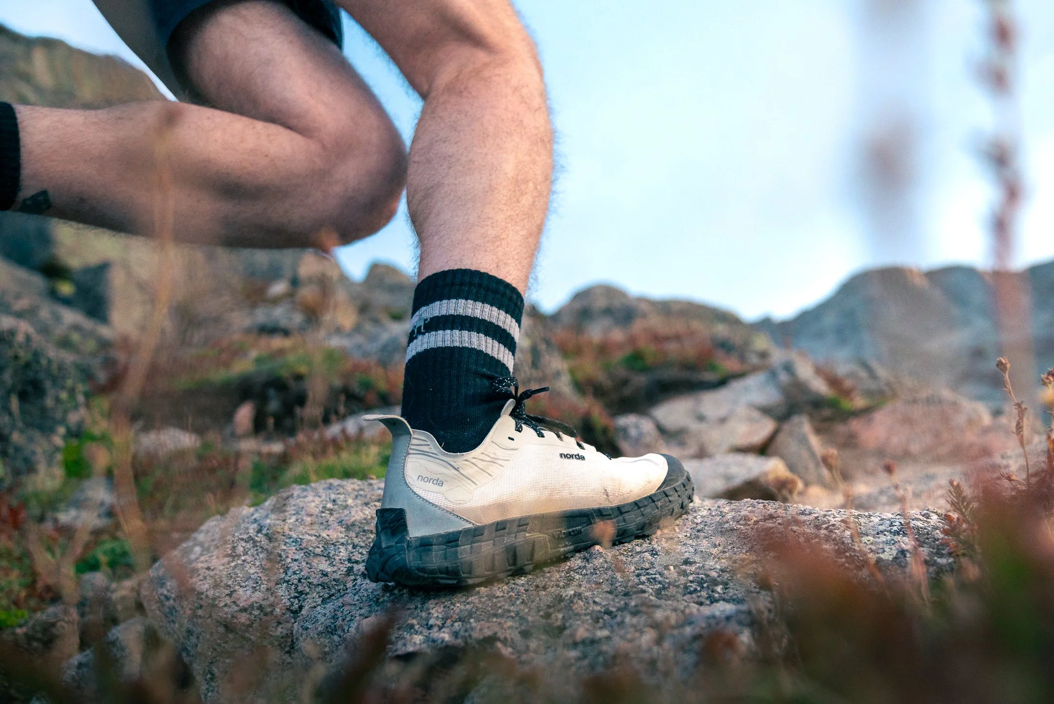 Close-up of a person wearing hiking shoes and black socks with gray stripes, standing on a rocky terrain outdoors with a blue sky background.