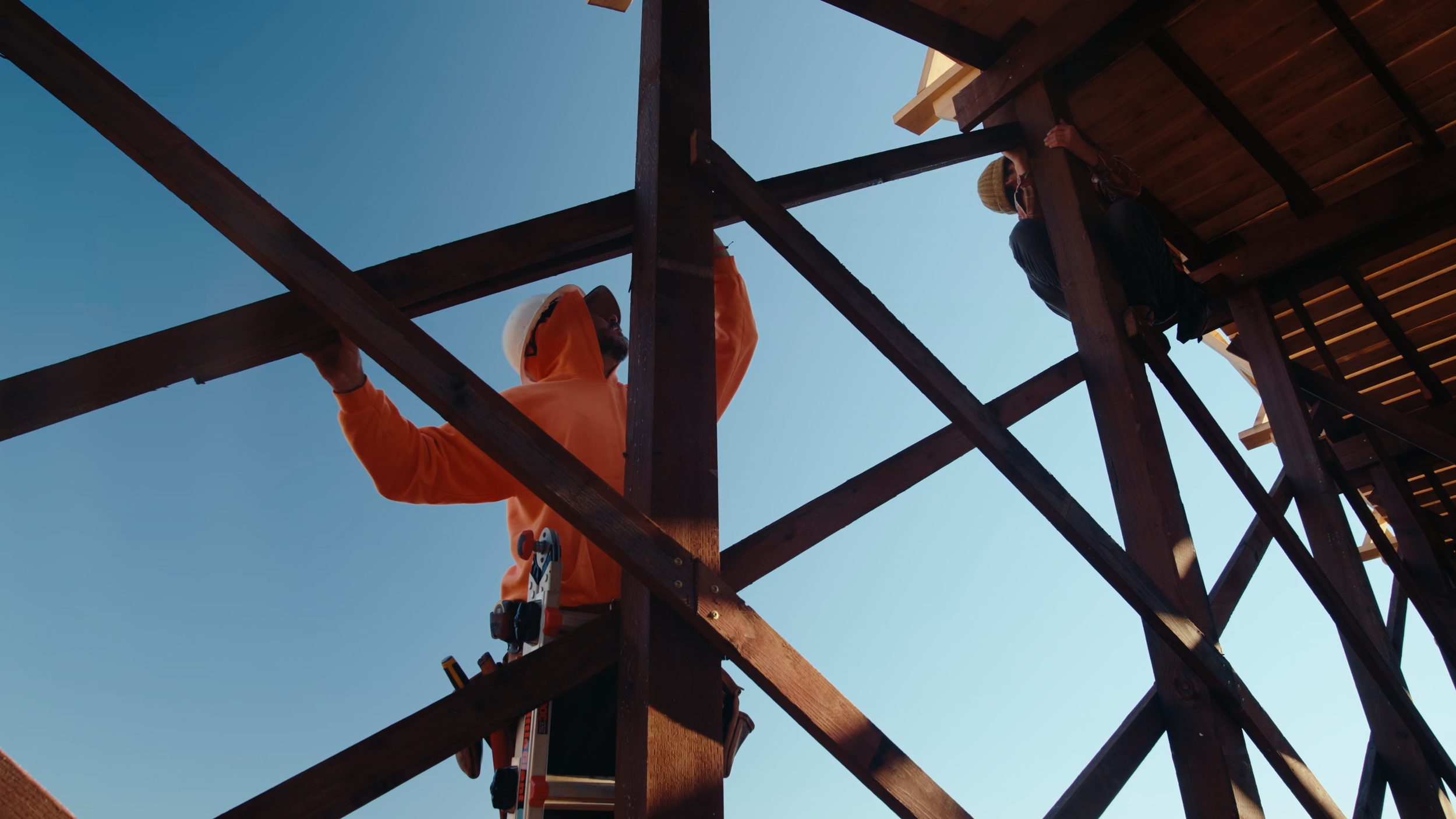 Construction workers building a wooden mountain bike ramp structure outdoors, viewed from below against a clear blue sky.