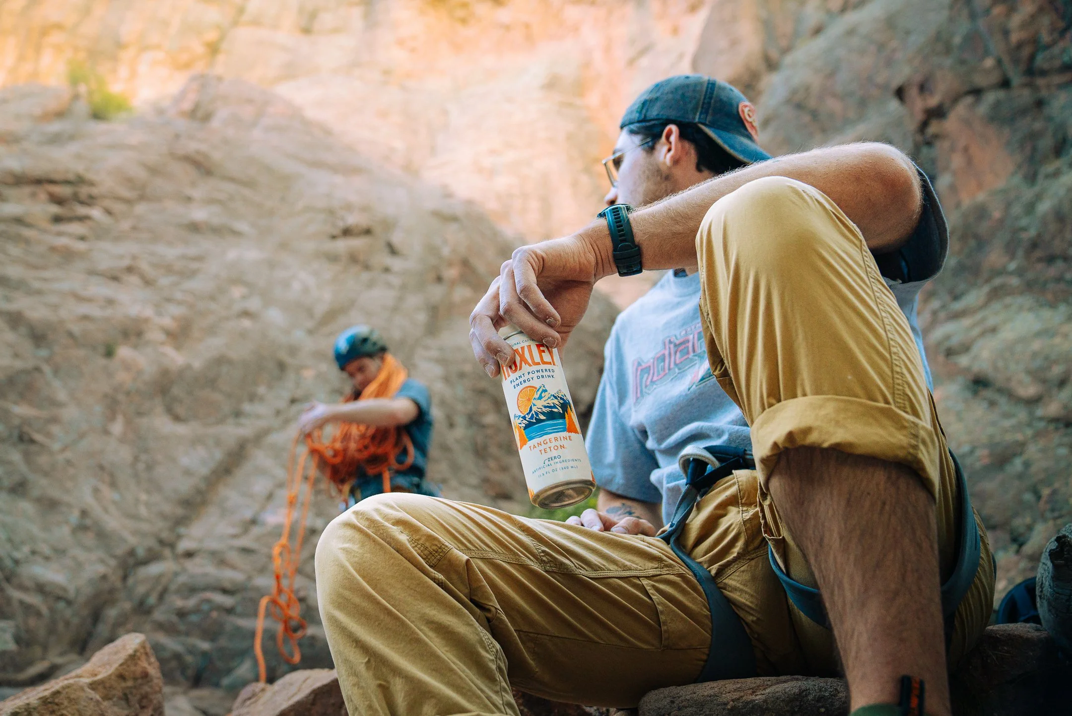 Two male climbers resting in a rocky outdoor setting, one sitting with a can of Endube energy drink, the other adjusting climbing gear in the background.