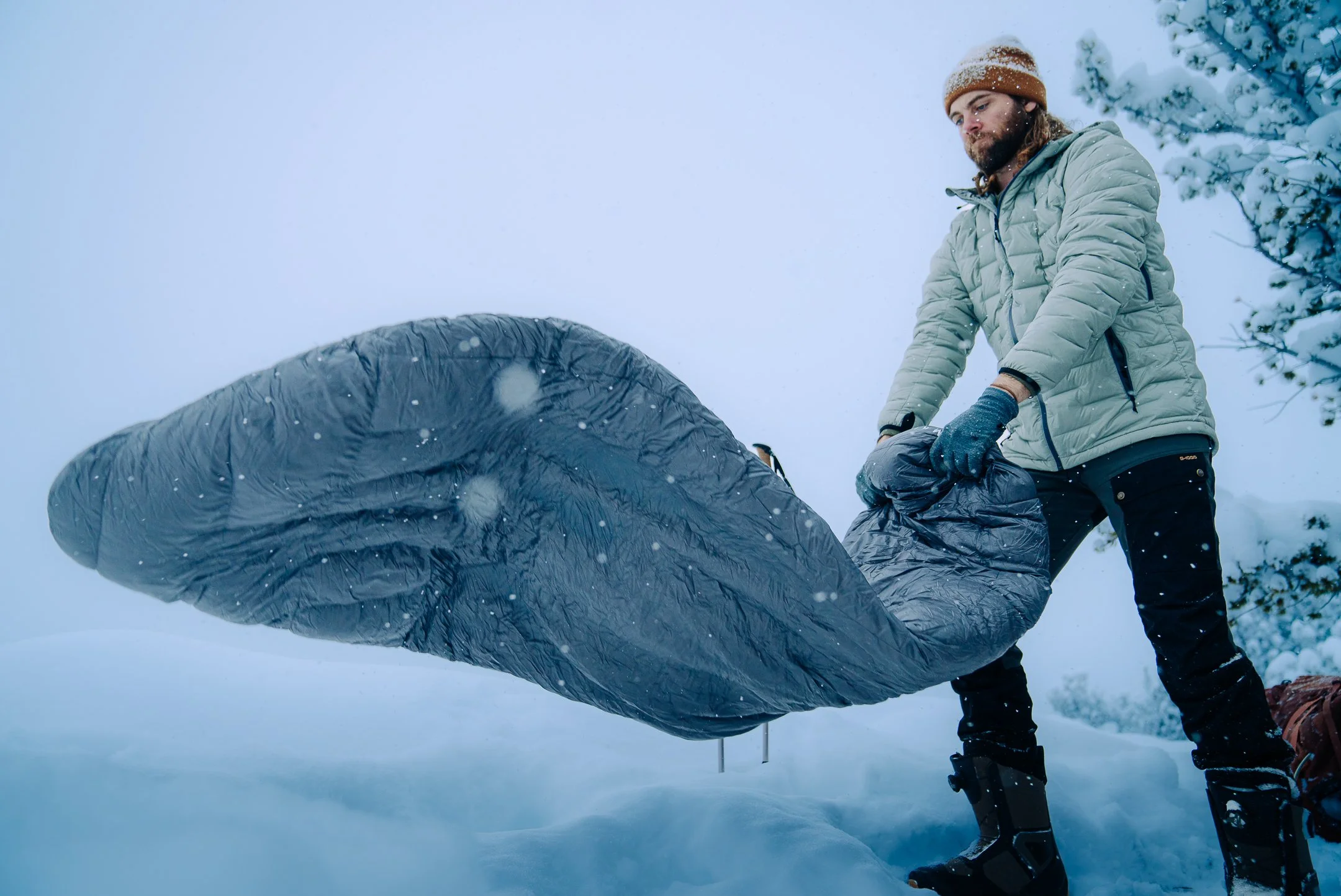 Man in winter jacket and gloves unfolding sleeping bag in snowy outdoors during snowstorm.