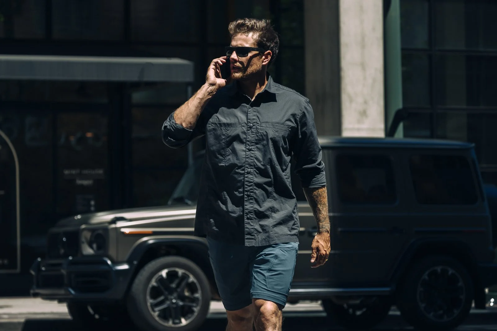 A man wearing sunglasses, a long-sleeved gray shirt, and shorts is walking while talking on a cellphone, with a black SUV parked behind him and a modern building in the background.