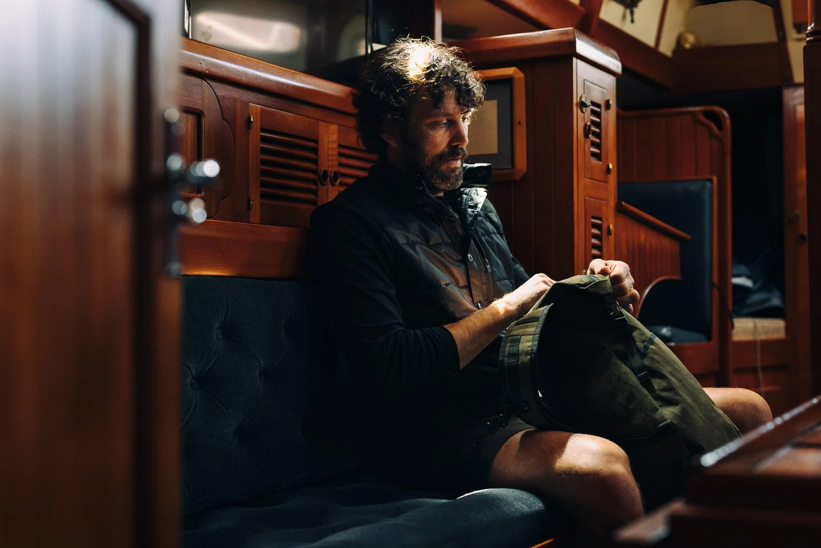 A man with dark curly hair and a beard sitting on a cushioned bench in a wooden interior, looking into a backpack.