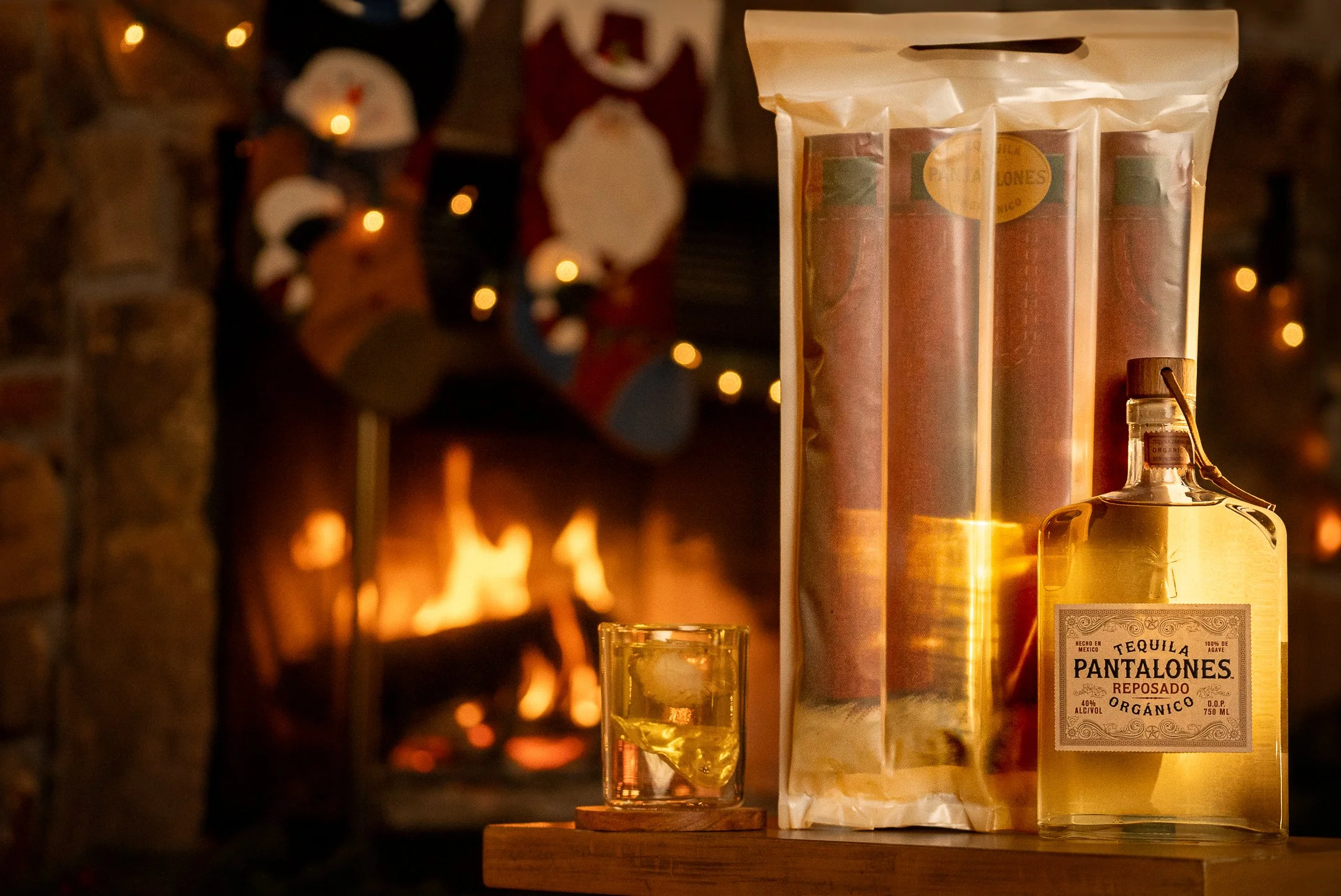 A bottle of Pantalones Reposado Organic tequila with a label, a glass of tequila with ice, and a package of rolled cigars on a wooden surface, with a lit fireplace and Christmas stockings hanging in the background.