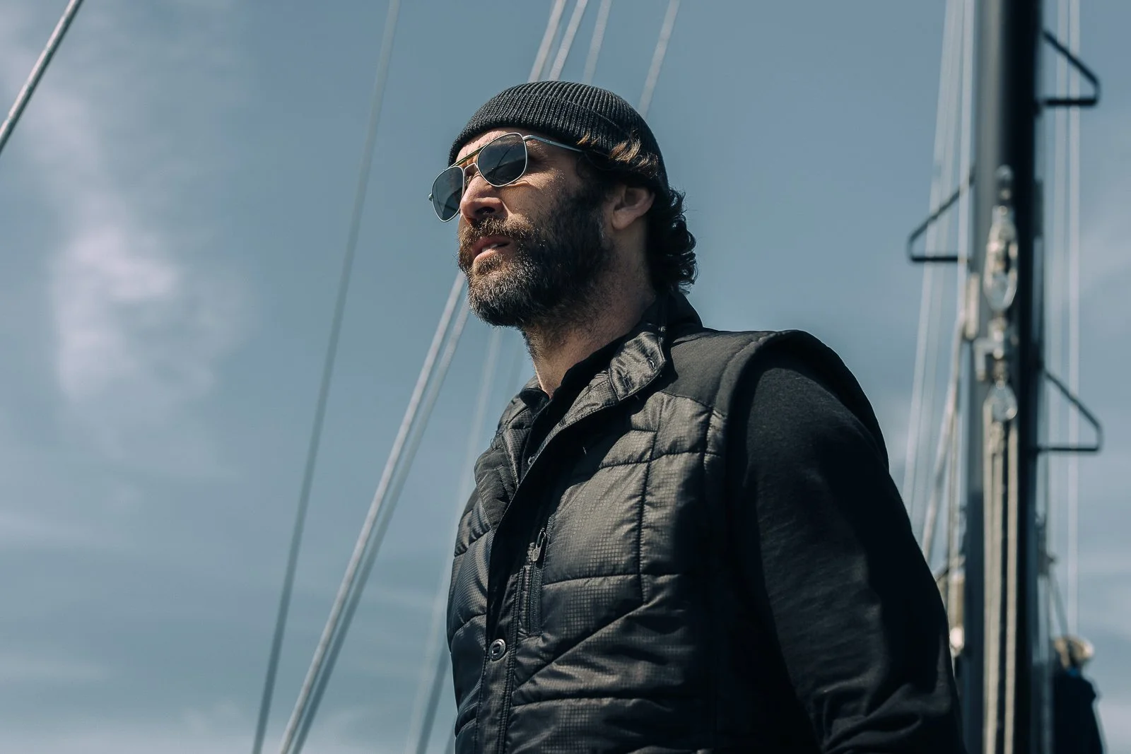 A man with sunglasses, a black beanie, a beard, and a black vest standing outdoors near a sailboat mast with ropes, against a cloudy sky.