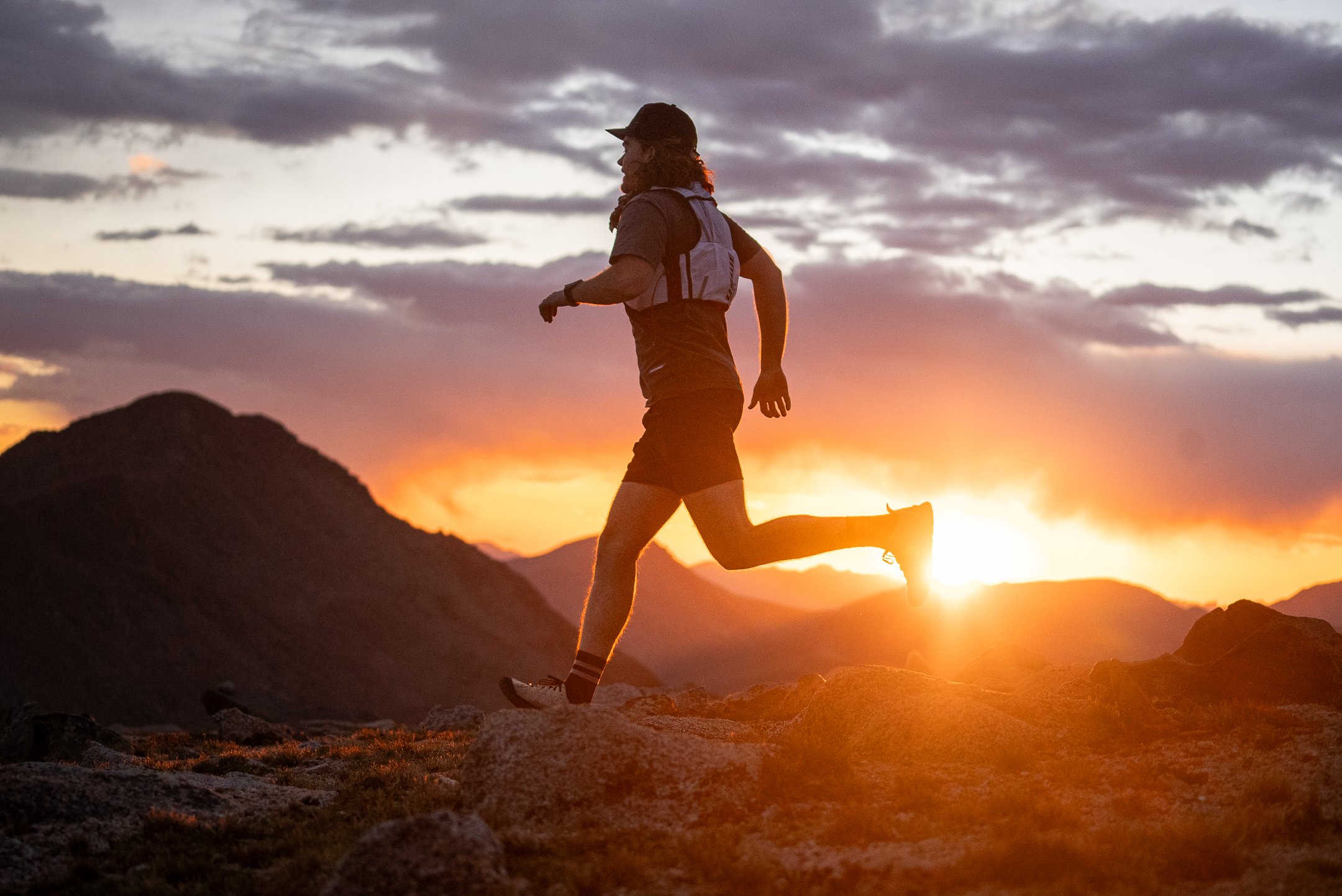 A person running on rocky terrain during sunset with mountains and a colorful sky in the background.