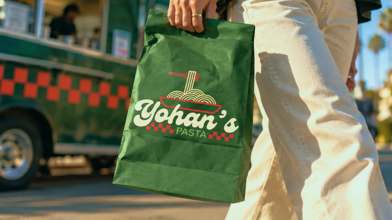 Person in white pants carrying a green Yohan's Pasta paper bag past a food truck